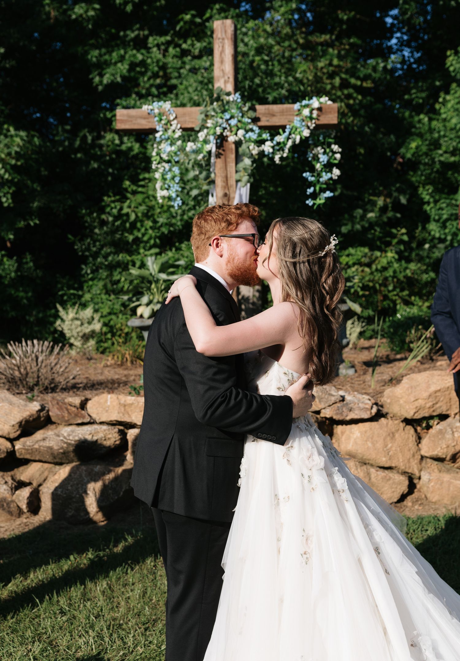 Bride and groom share first kiss at outdoor wedding ceremony with wooden cross decorated with greenery.