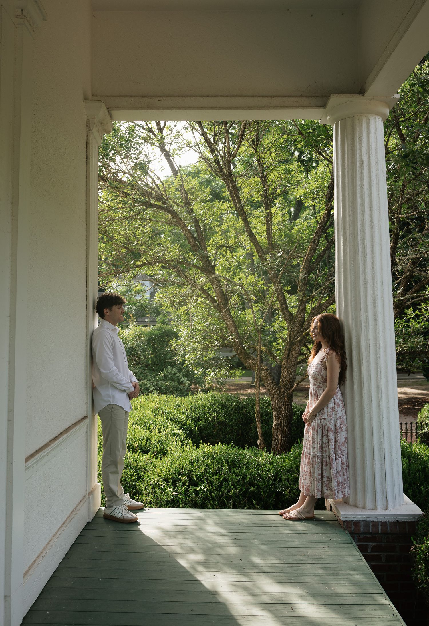 A young couple stands on a sunlit porch between white columns, surrounded by lush green gardens.