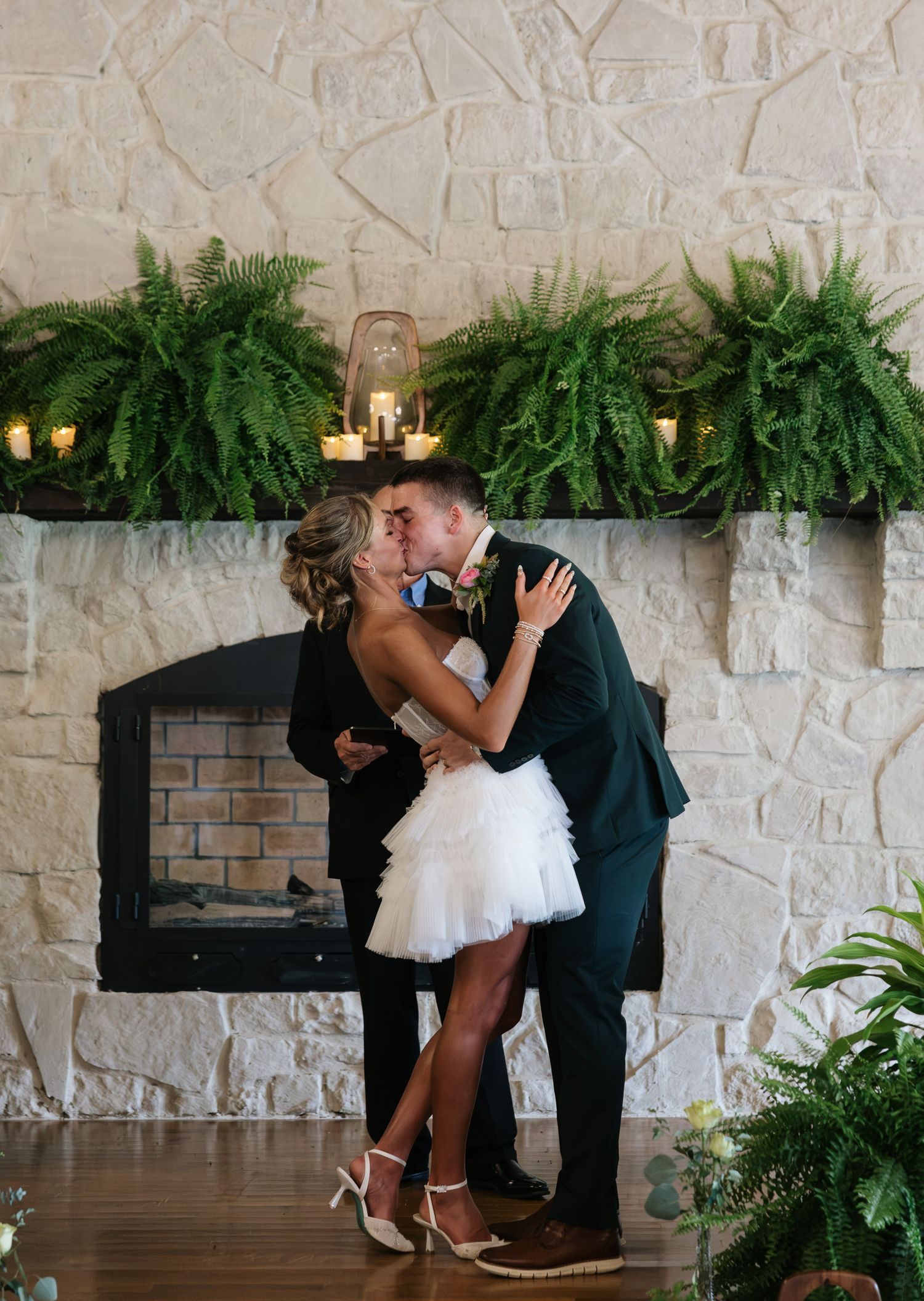 Newlyweds share a first kiss in front of a stone fireplace adorned with lush fern garland and candles.
