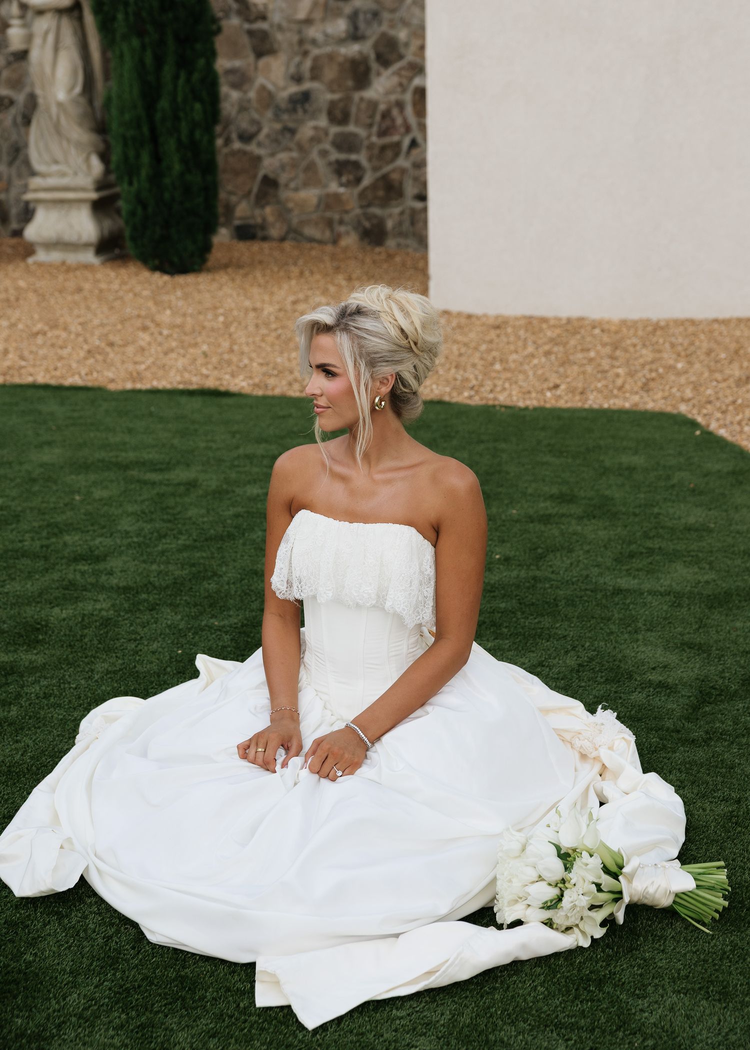 Bride in strapless white ball gown sitting on green lawn holding white floral bouquet at outdoor wedding venue.