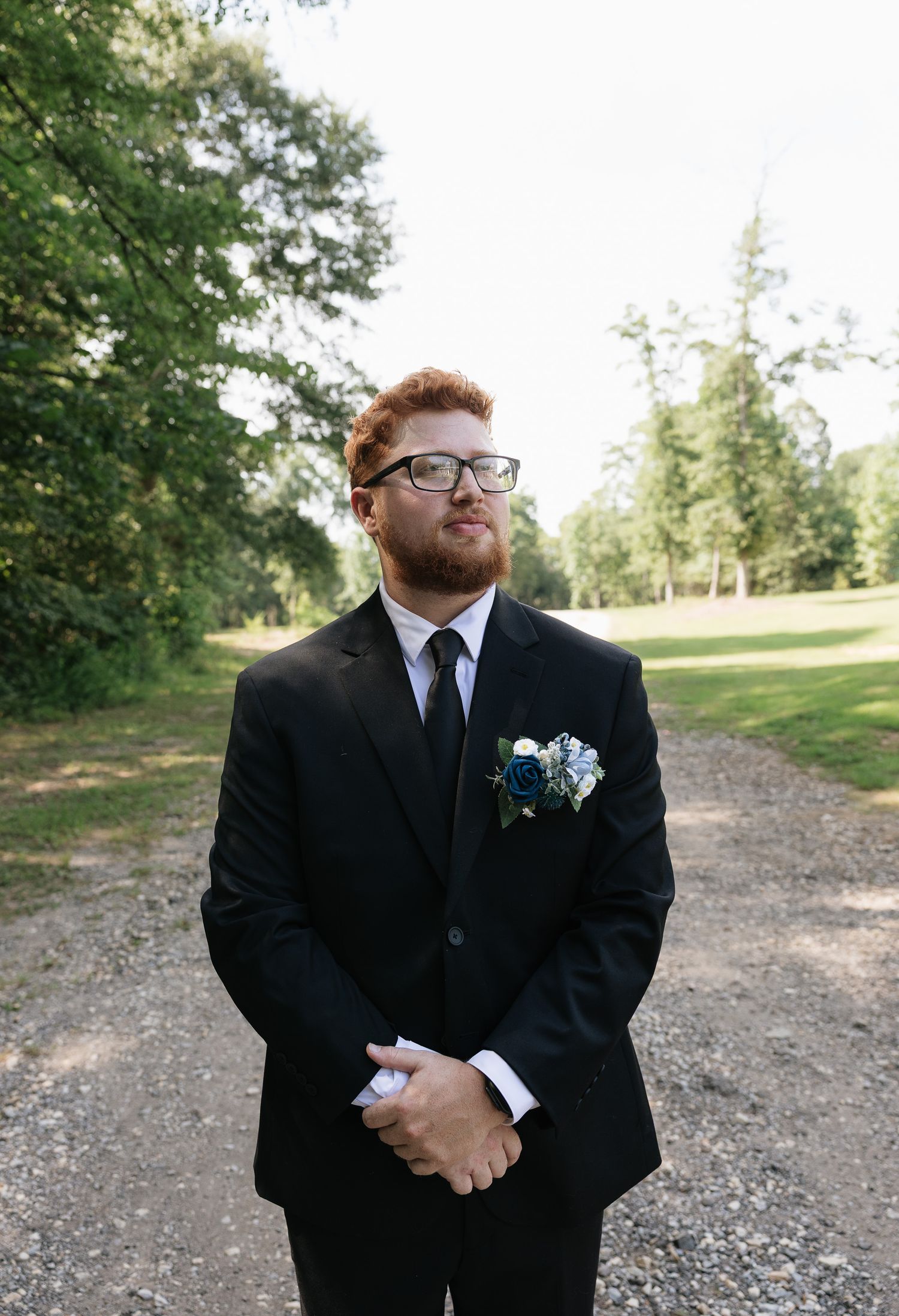 Groom in black suit with blue floral boutonniere standing on gravel path surrounded by green trees outdoors.