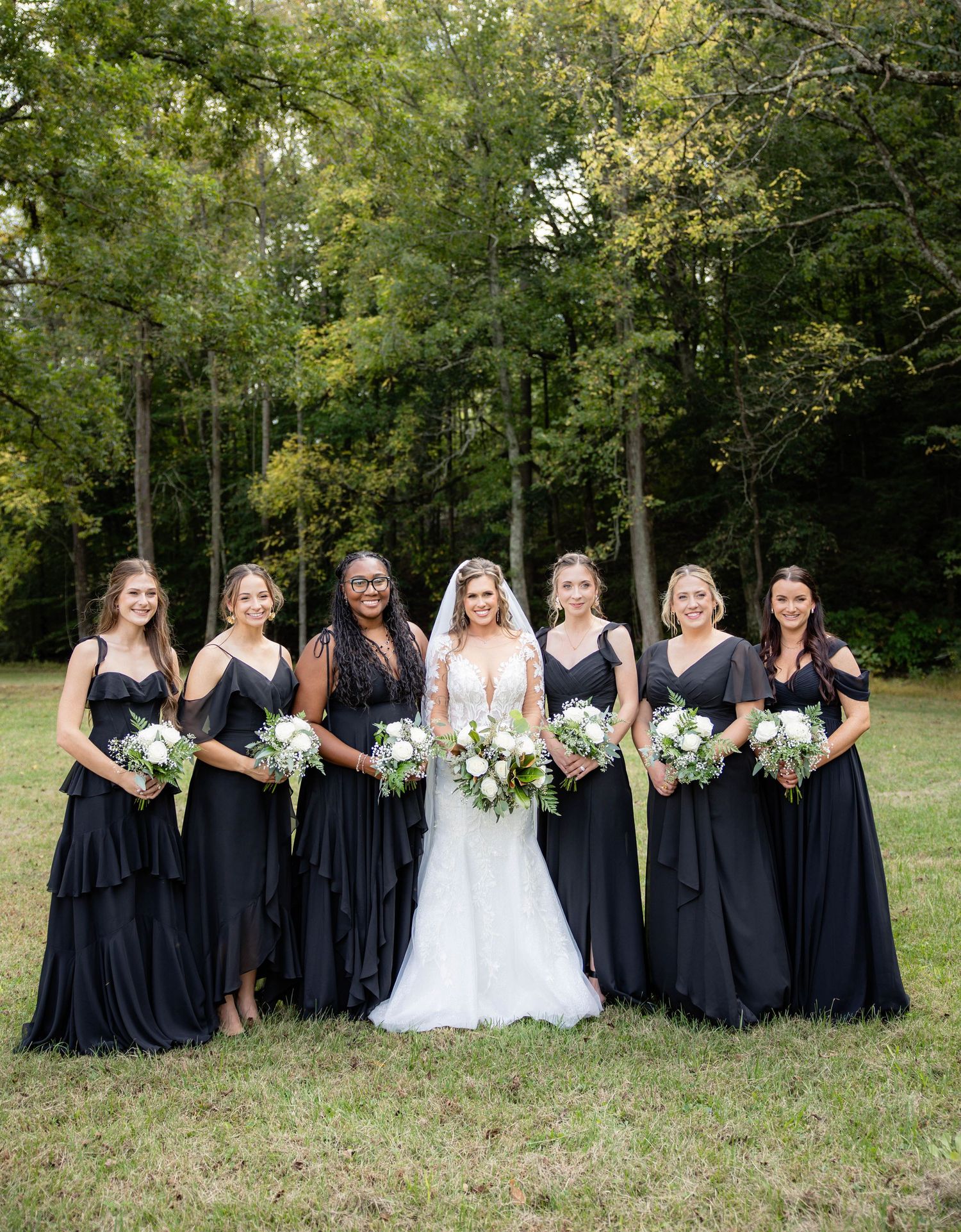 Wedding party in navy dresses poses together on grass with trees in background.