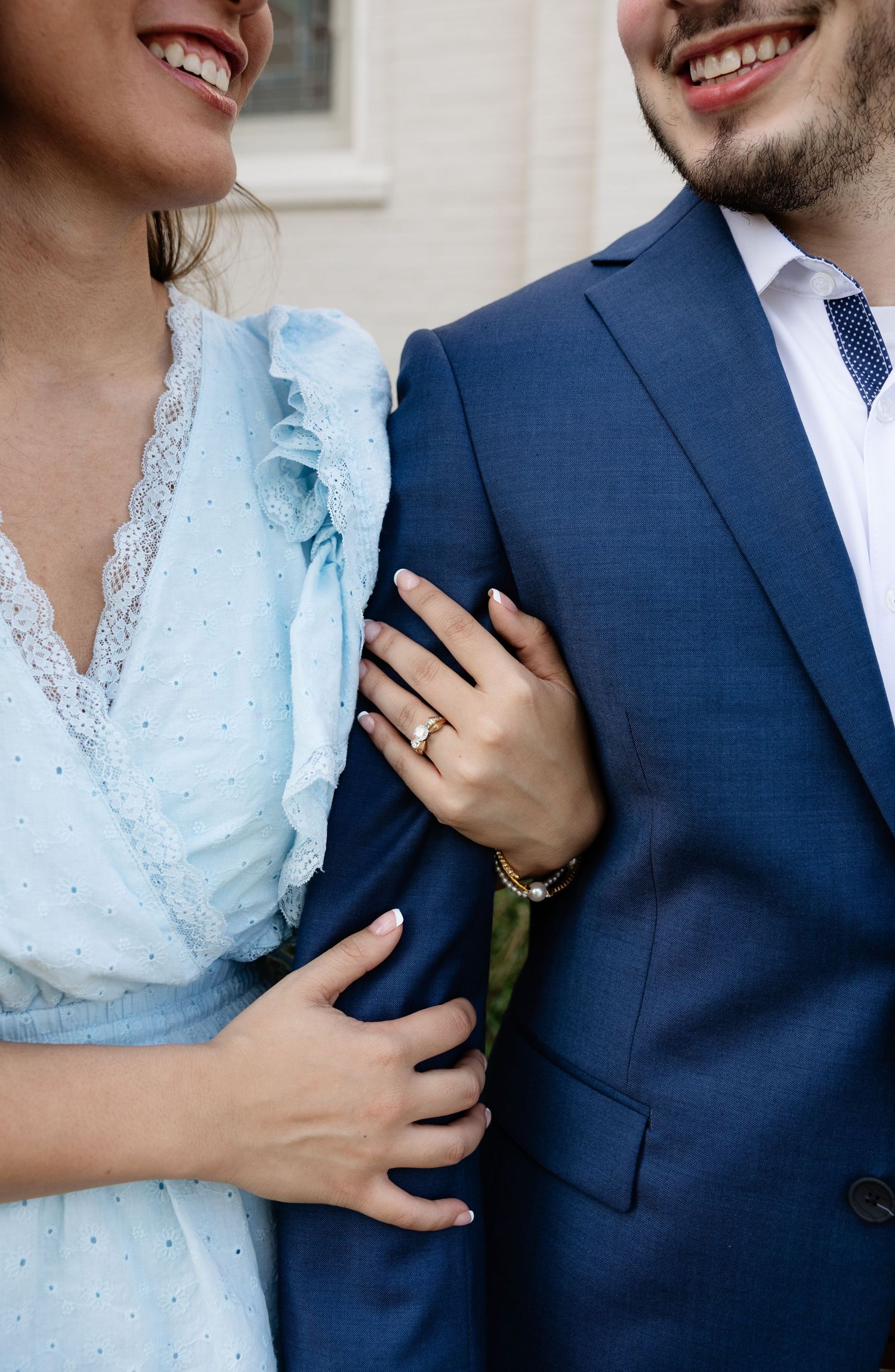 Smiling couple posing together, woman in blue lace dress with gold ring holds man's arm in navy suit.