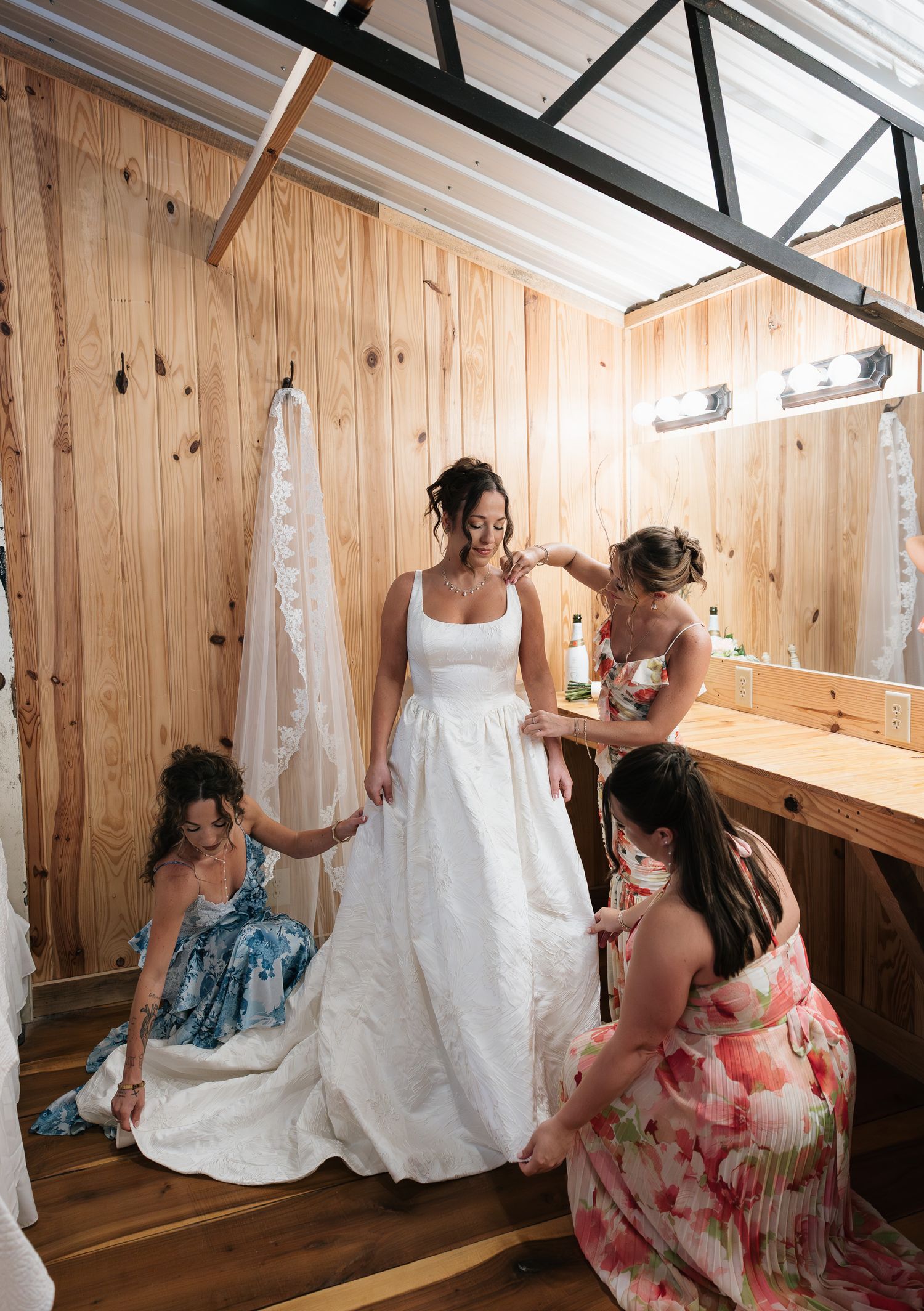 Bride in white gown being helped by bridesmaids in floral dresses inside a rustic wooden bridal suite.