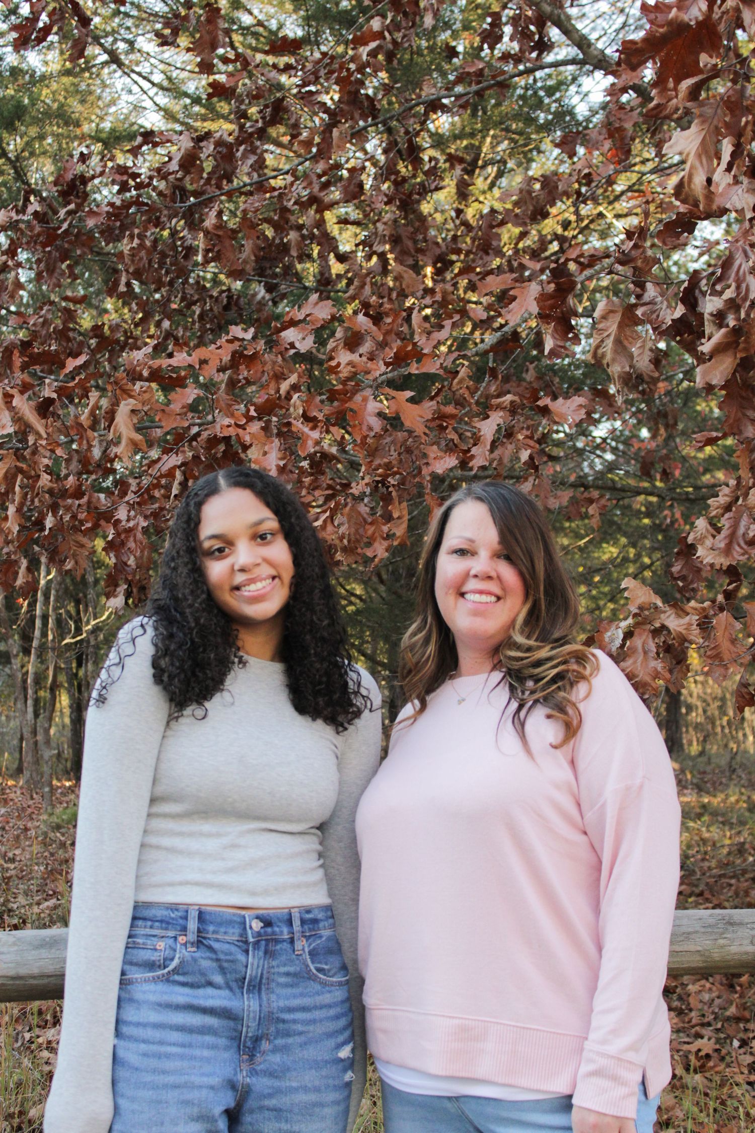 Two friends in pink and cream sweaters pose together against a backdrop of fall trees.