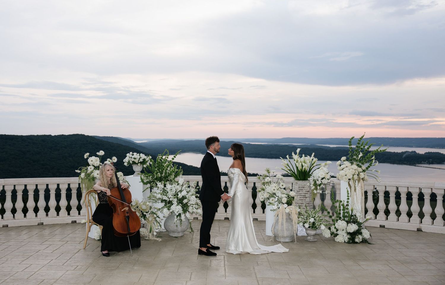 Elegant outdoor wedding ceremony on a terrace with white floral arrangements, a cellist, and scenic river views at dusk.