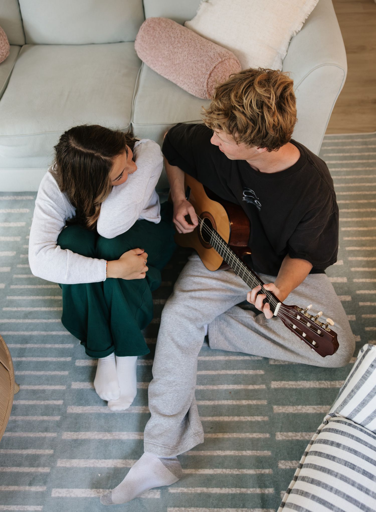 Couple sitting on floor against sofa, young man playing acoustic guitar while woman listens and smiles.