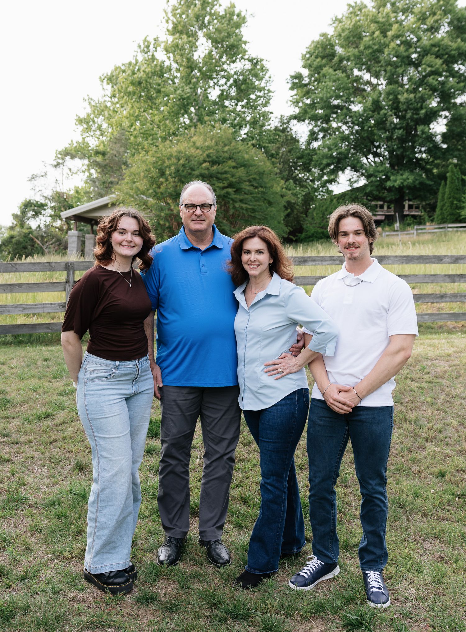 Family of four smiling together outdoors on a green lawn near a wooden fence on a sunny day.