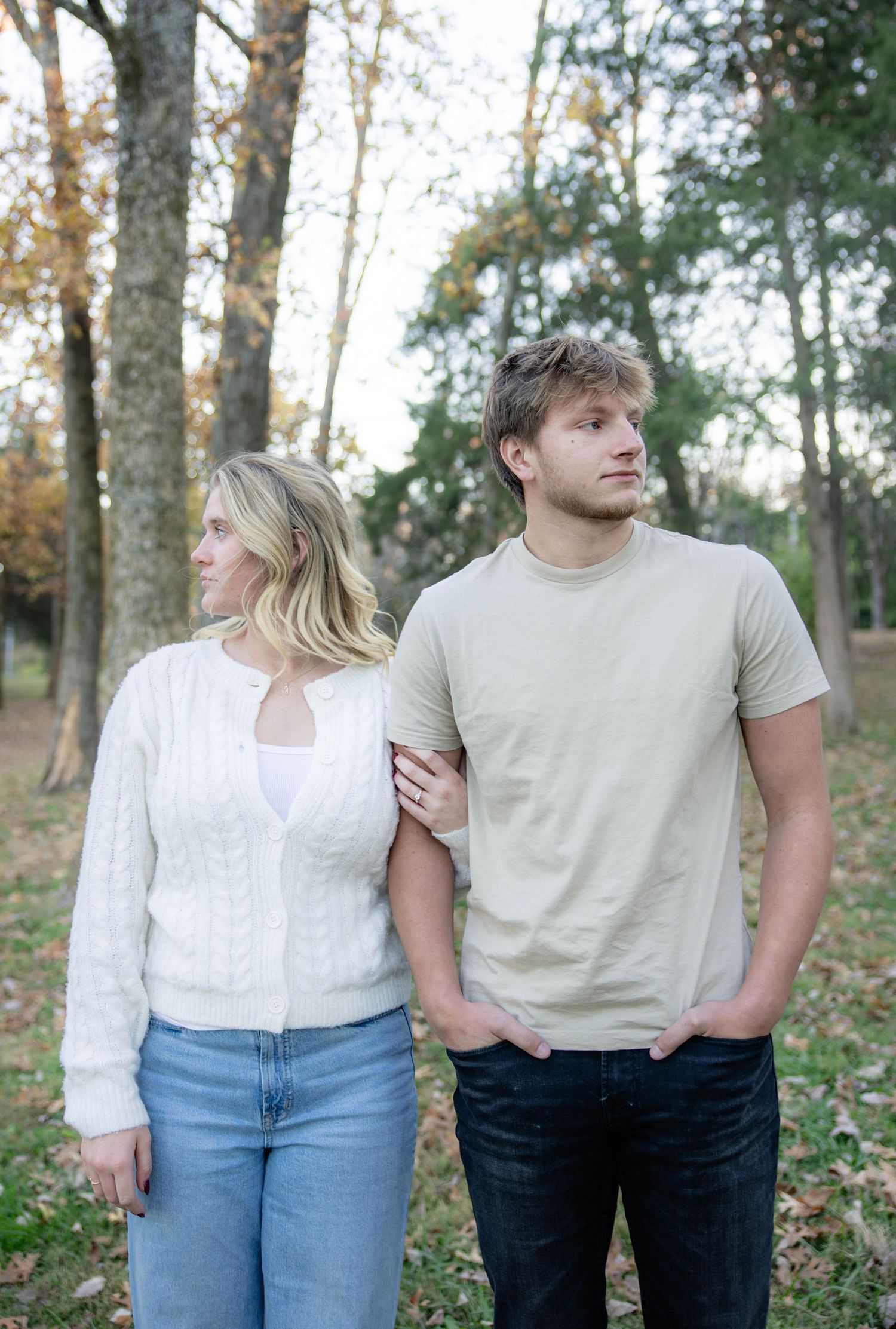 A couple in casual outfits stands together in a wooded area with autumn trees in the background.