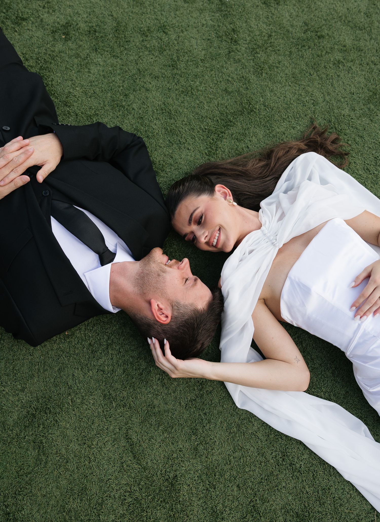 Newlyweds lying on green grass smiling at each other in wedding attire during outdoor photo session.
