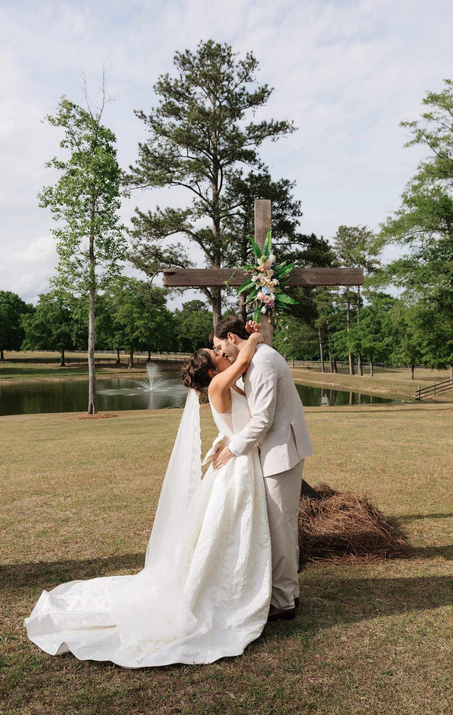 Bride and groom sharing a kiss in front of a floral cross near a pond on their outdoor wedding day.