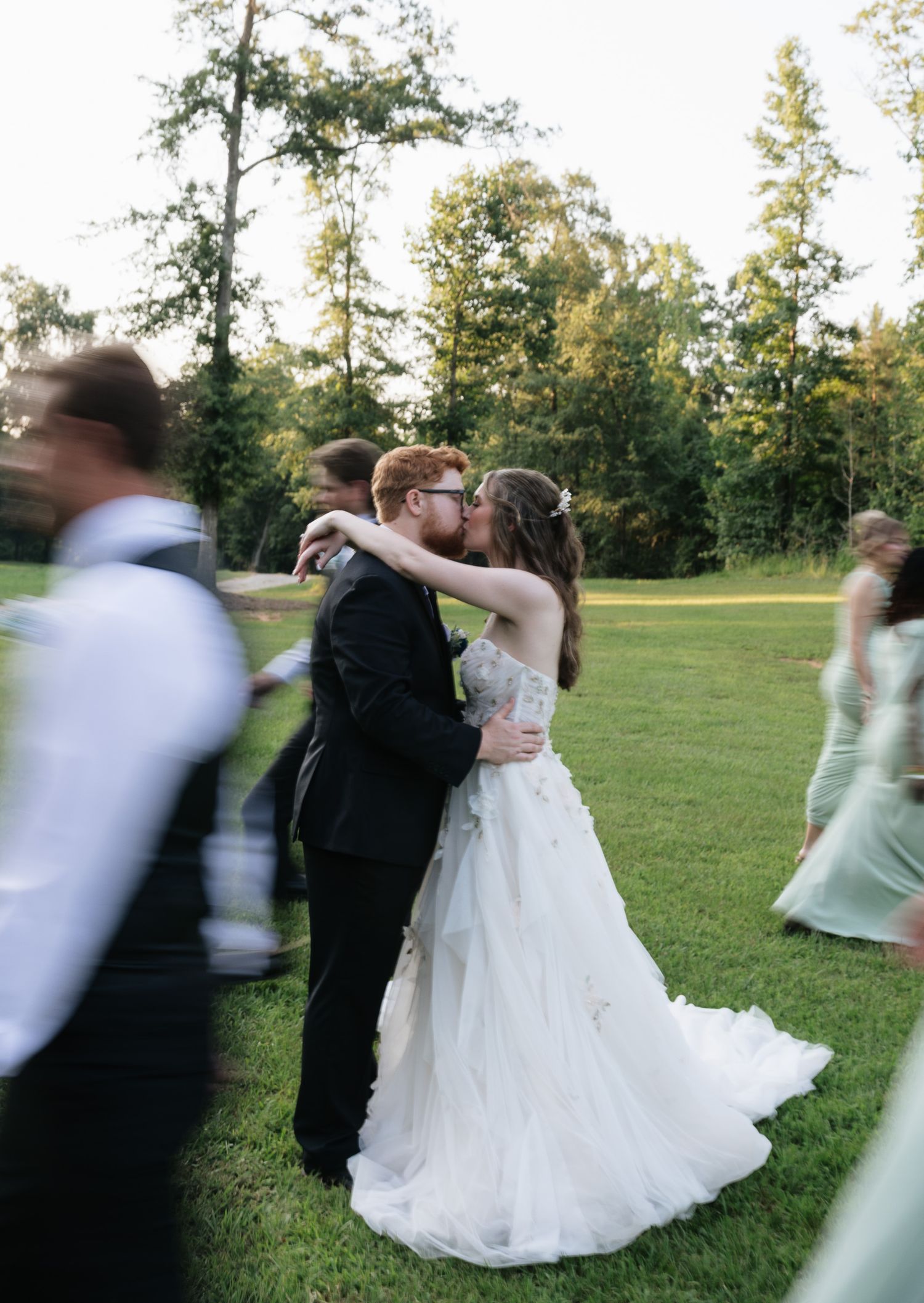 A bride and groom share a kiss outdoors on a green lawn, surrounded by wedding guests, with tall trees in the background.