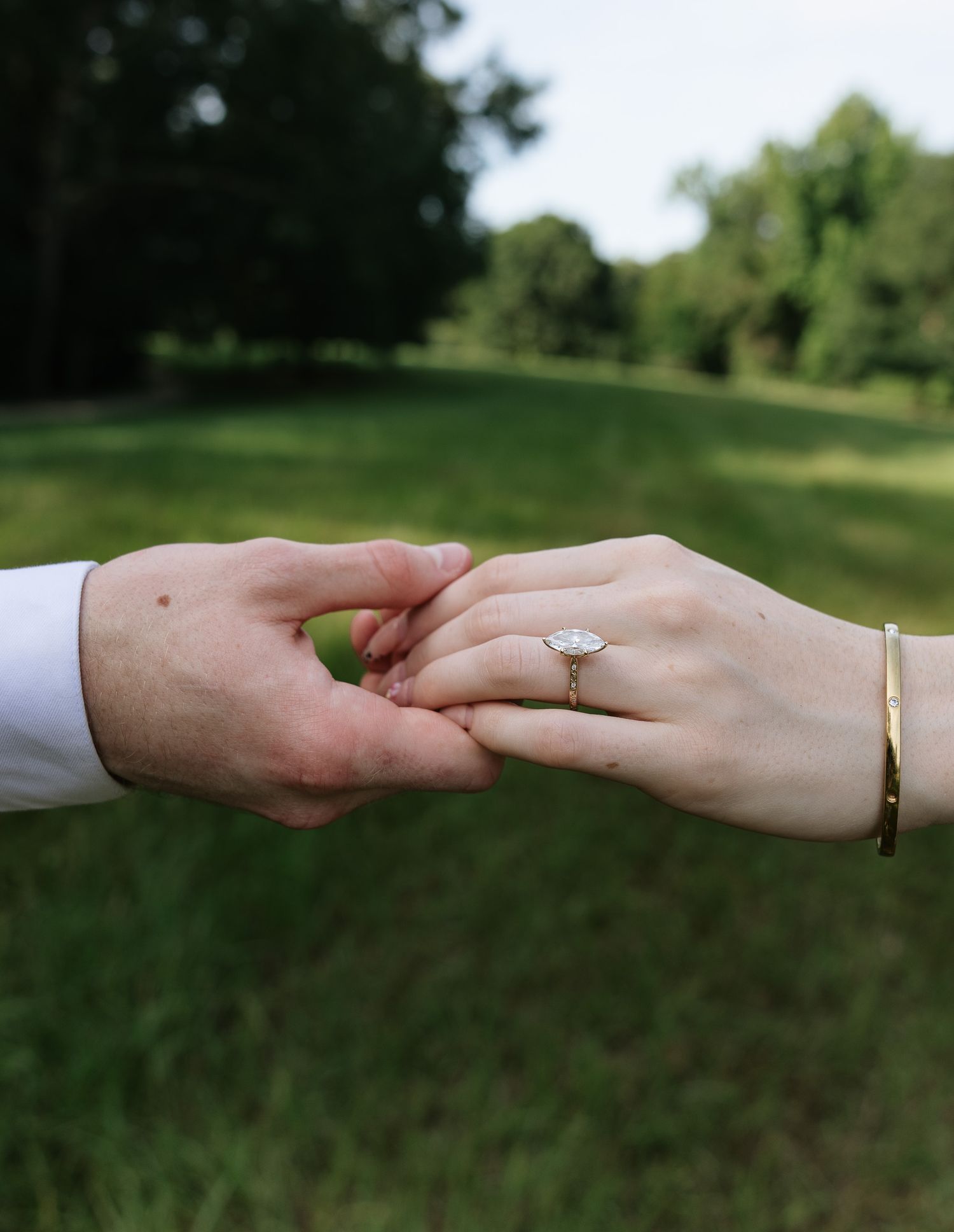 Two hands gently touching outdoors, woman wearing a diamond engagement ring and gold bangle bracelet on a green lawn.