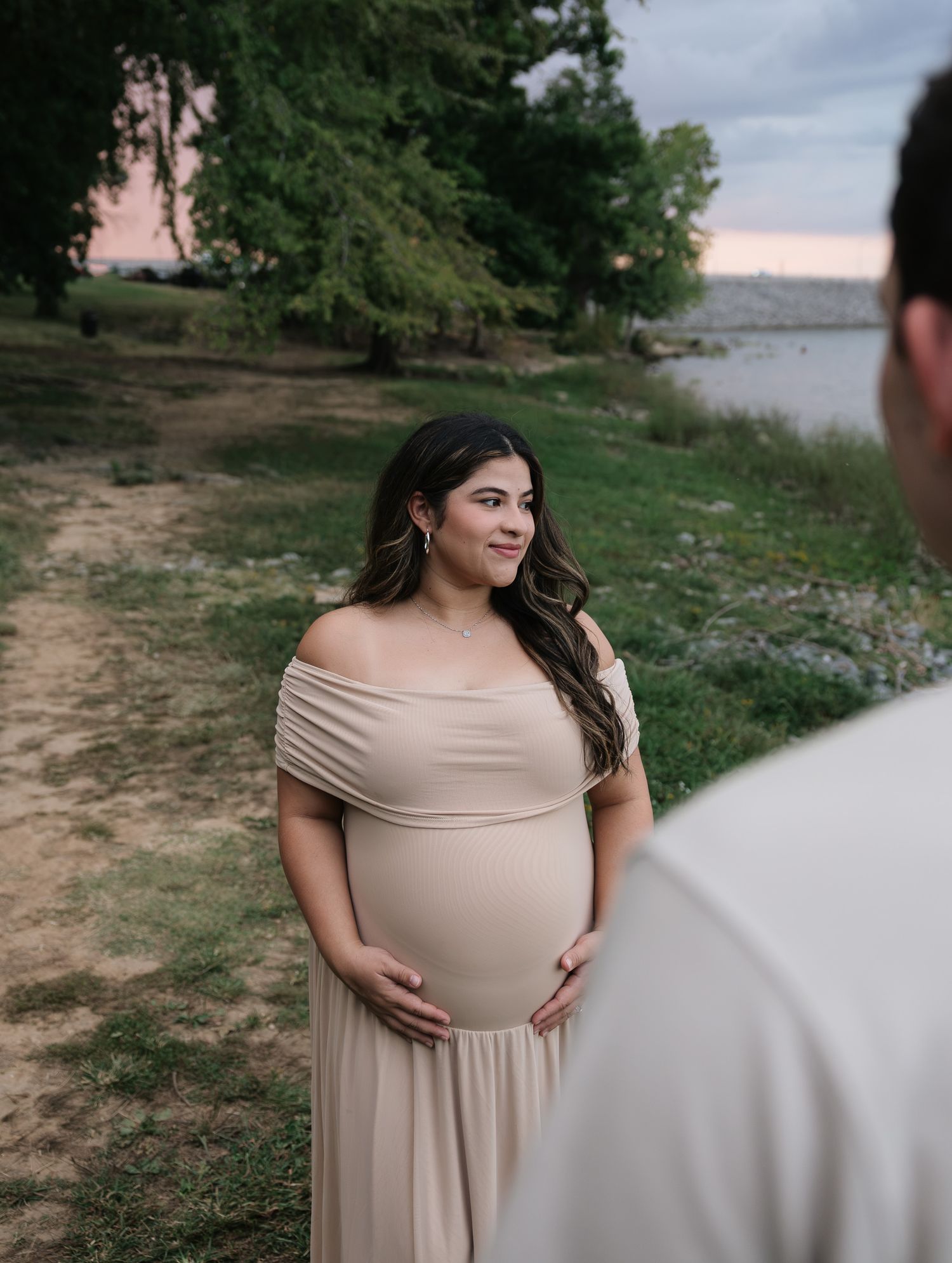 Pregnant woman in beige off-shoulder gown cradles baby bump near lakeside at dusk during maternity photoshoot.