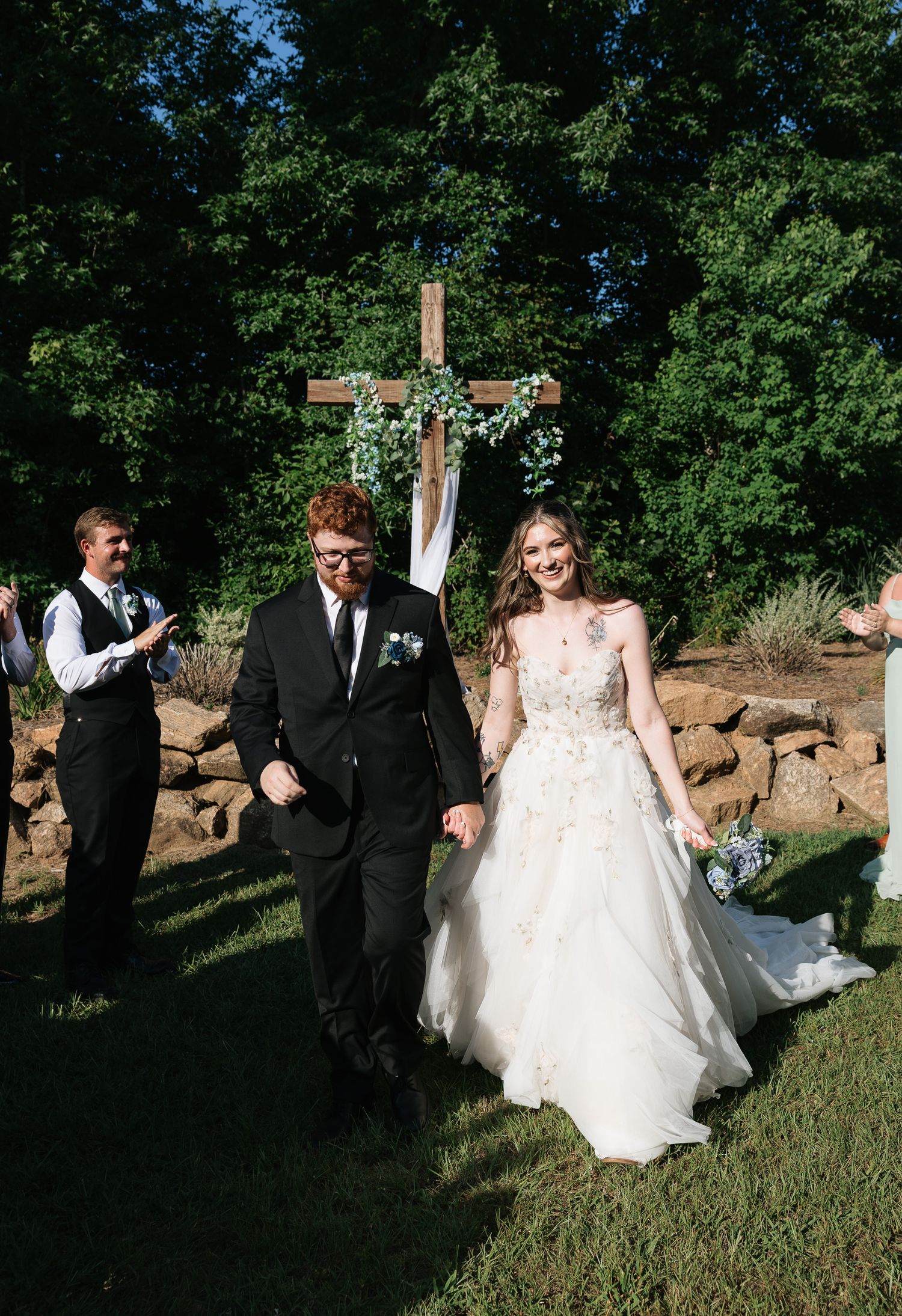 Newlyweds walk back down the aisle smiling at outdoor wedding ceremony with wooden cross backdrop.