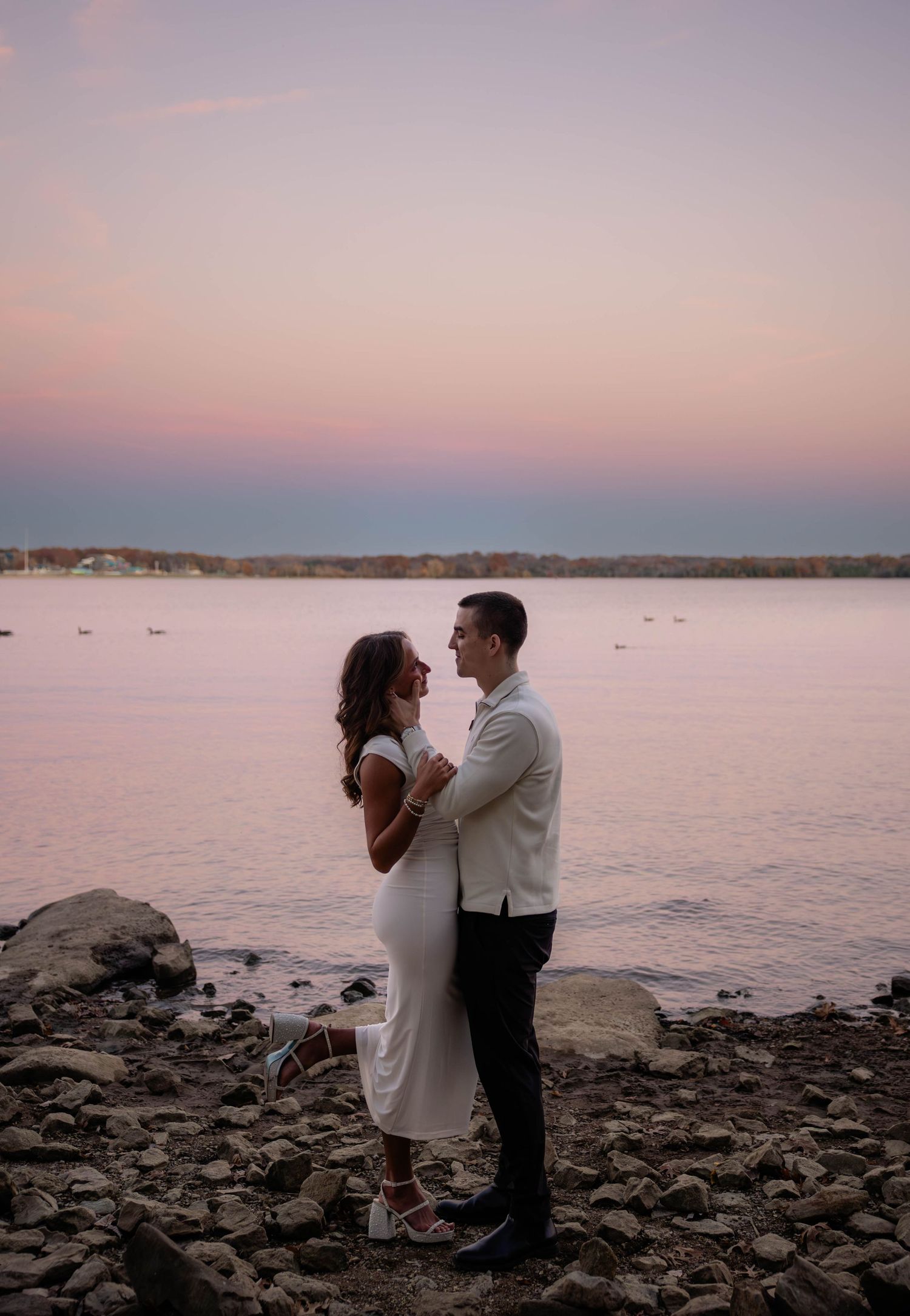 A couple shares a romantic moment at sunset on a rocky beach overlooking calm waters.