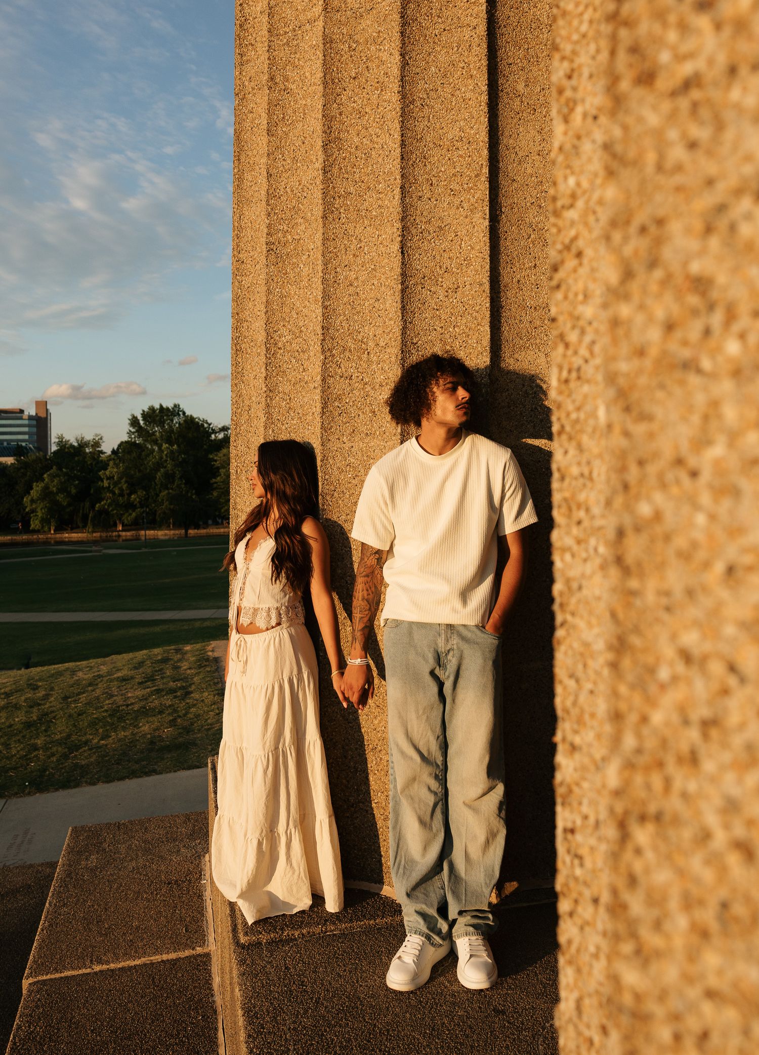 A couple holds hands between large stone columns at golden hour, she in a white maxi dress, he in a white tee and gray pants.