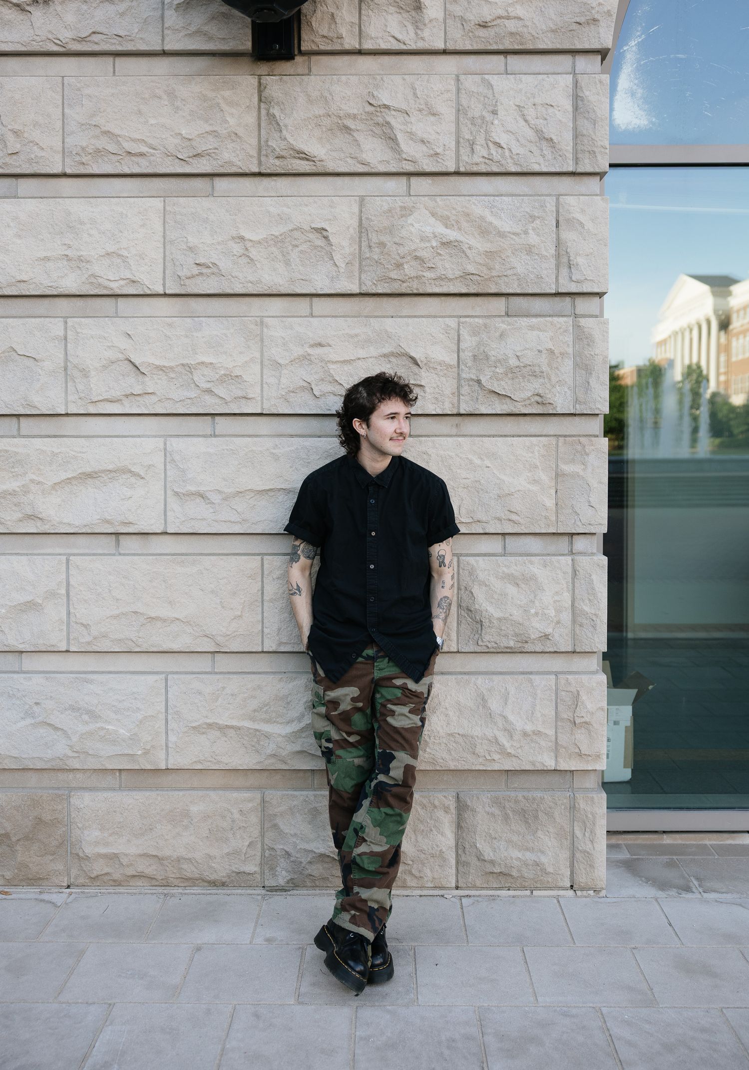 Young man in black shirt and camo pants leaning against a stone wall outdoors, smiling and looking away.