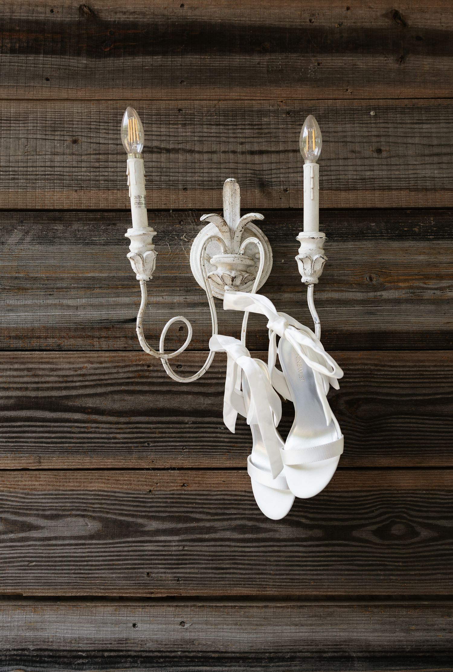 White bridal heels hanging from a vintage white candle wall sconce against a rustic wood plank wall.