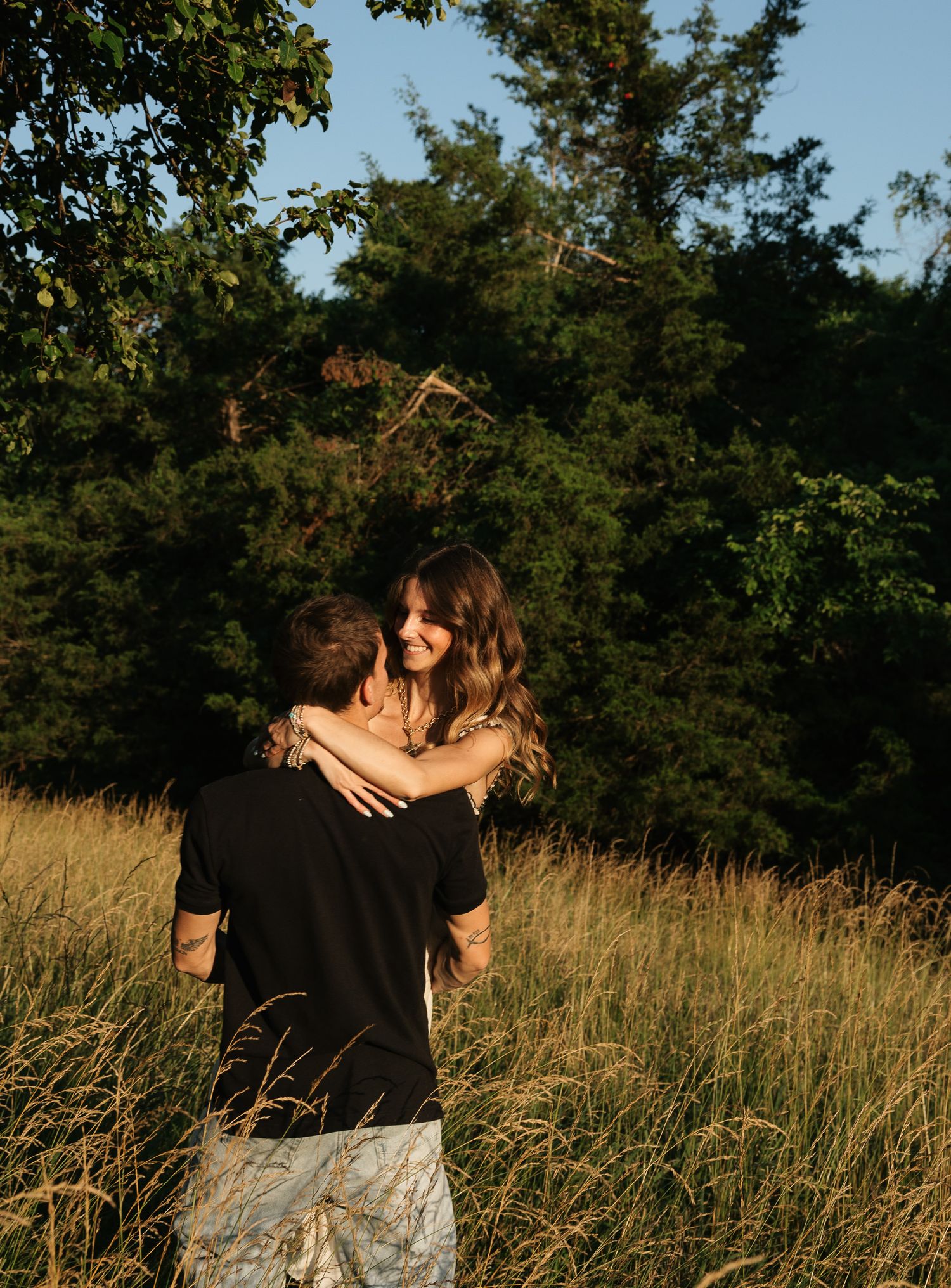 A couple embraces in a golden field surrounded by lush green trees during a warm sunset outdoor photo session.
