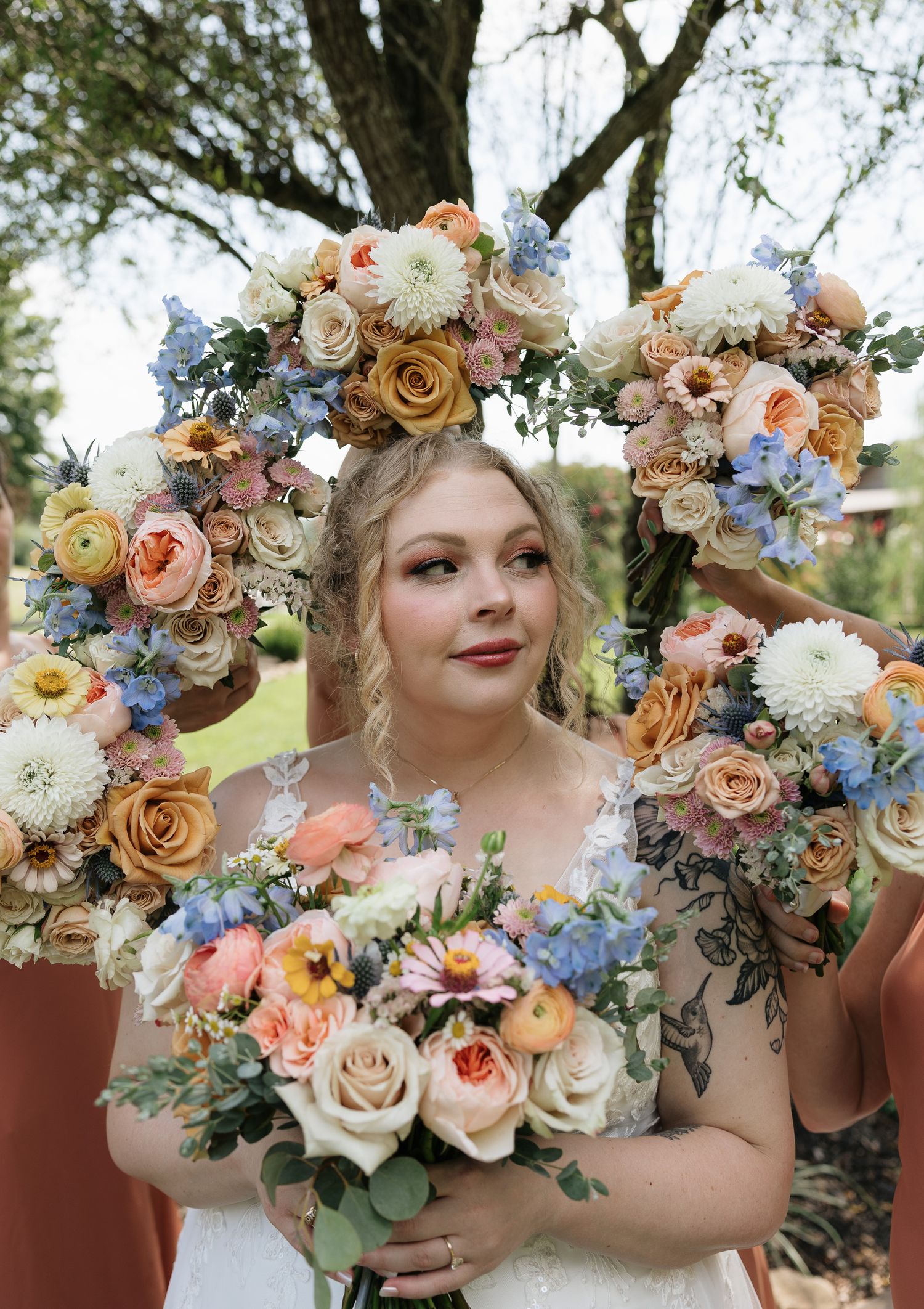 Bride holding colorful floral bouquet framed by floral letter decoration with peach, blue, and cream roses outdoors.
