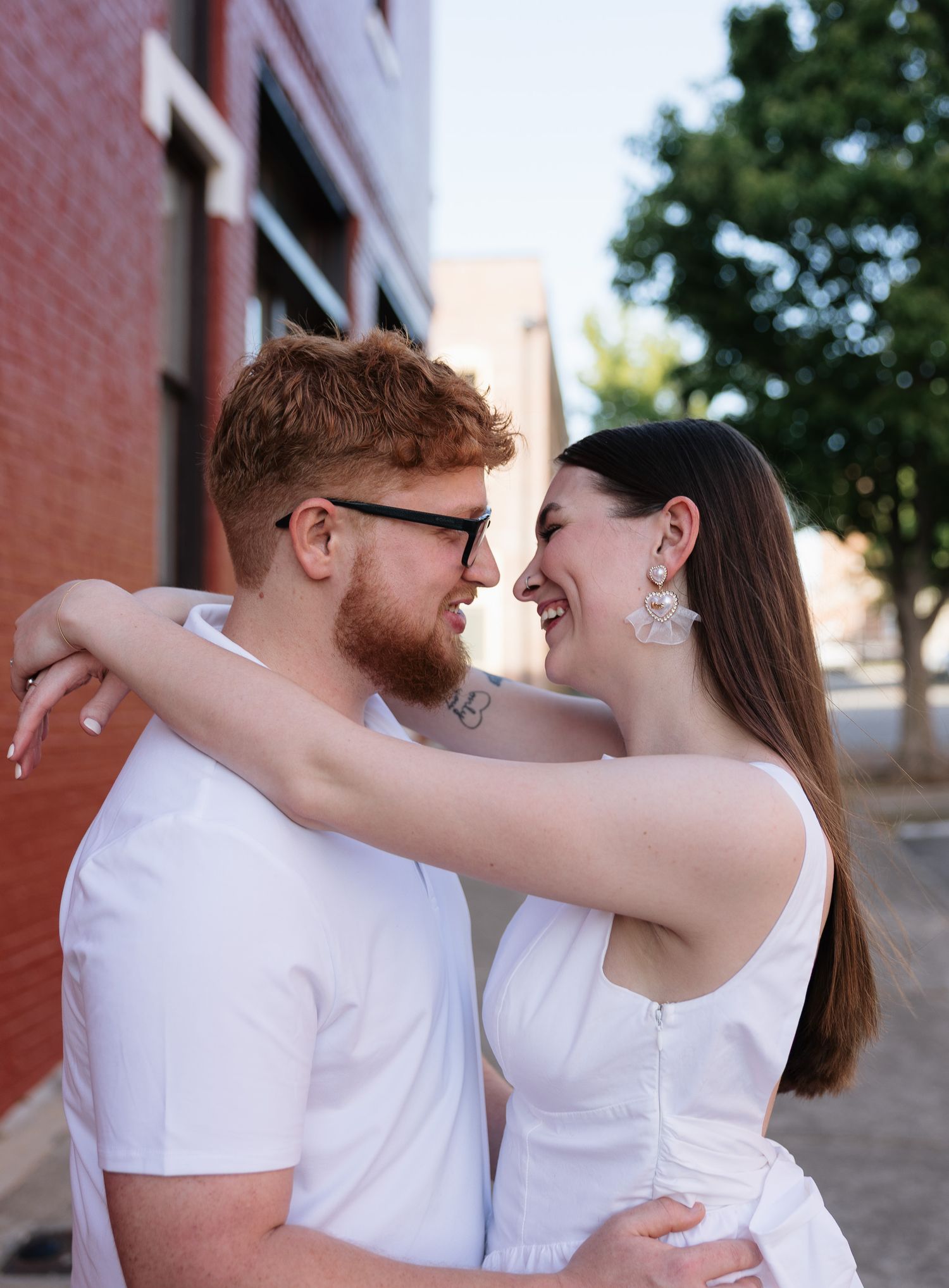 A smiling couple in matching white outfits share a romantic moment outdoors near a brick building on a sunny day.