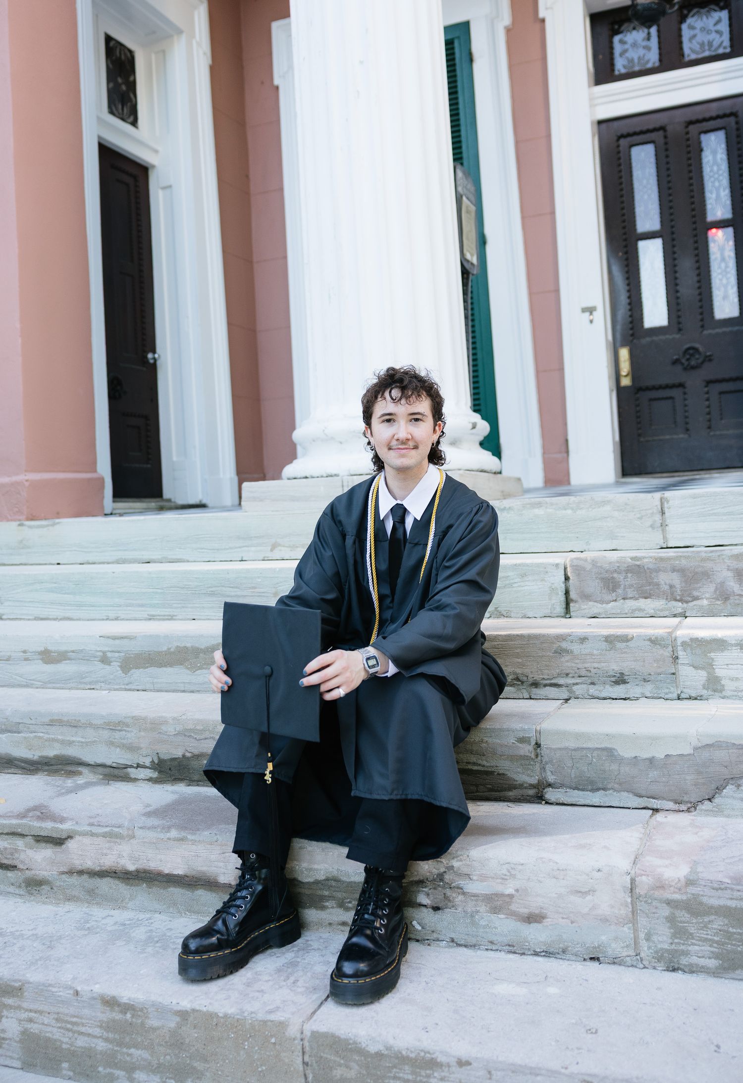 A smiling graduate in black cap and gown sits on stone steps holding their mortarboard, wearing gold honor cords.