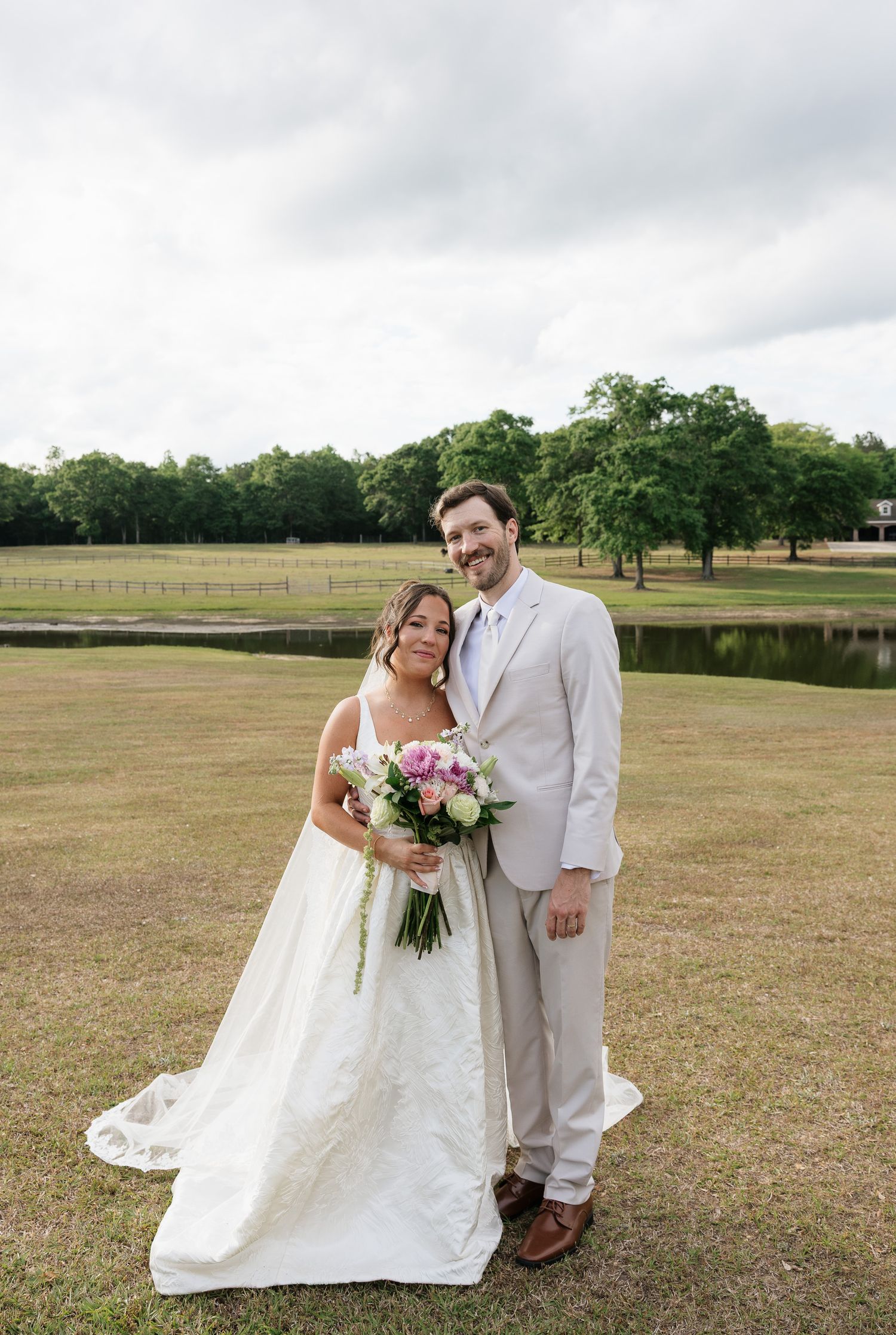 Bride in white gown holding pink bouquet stands with groom in tan suit on green lawn near pond.