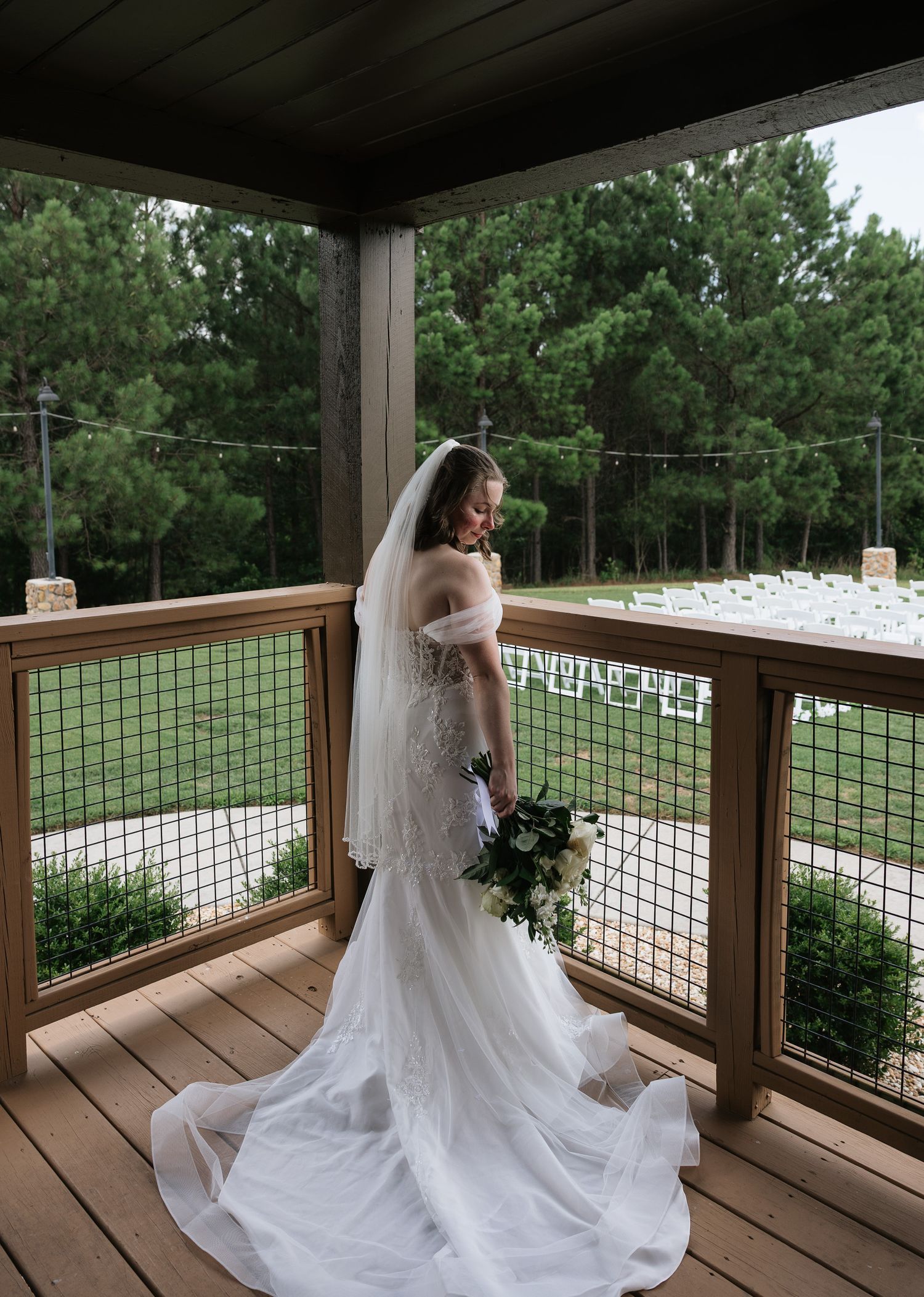 Bride in white off-shoulder gown with veil and bouquet stands on wooden deck overlooking outdoor wedding ceremony setup.