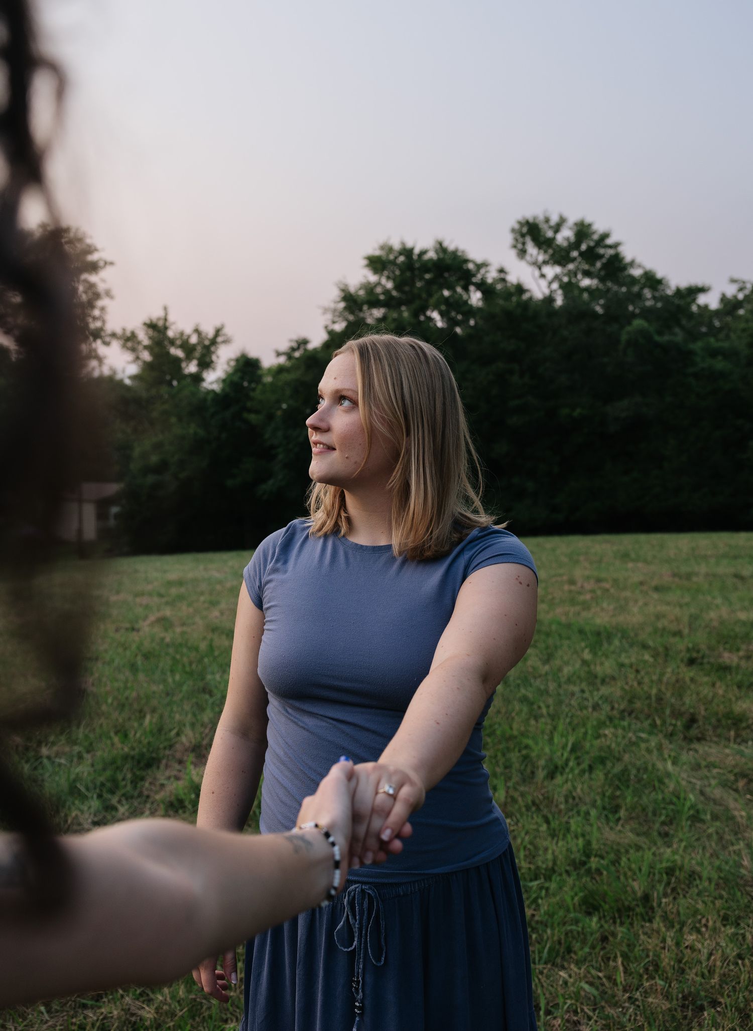 Woman in blue dress holding hands in a green field at dusk, looking up thoughtfully with trees in background.