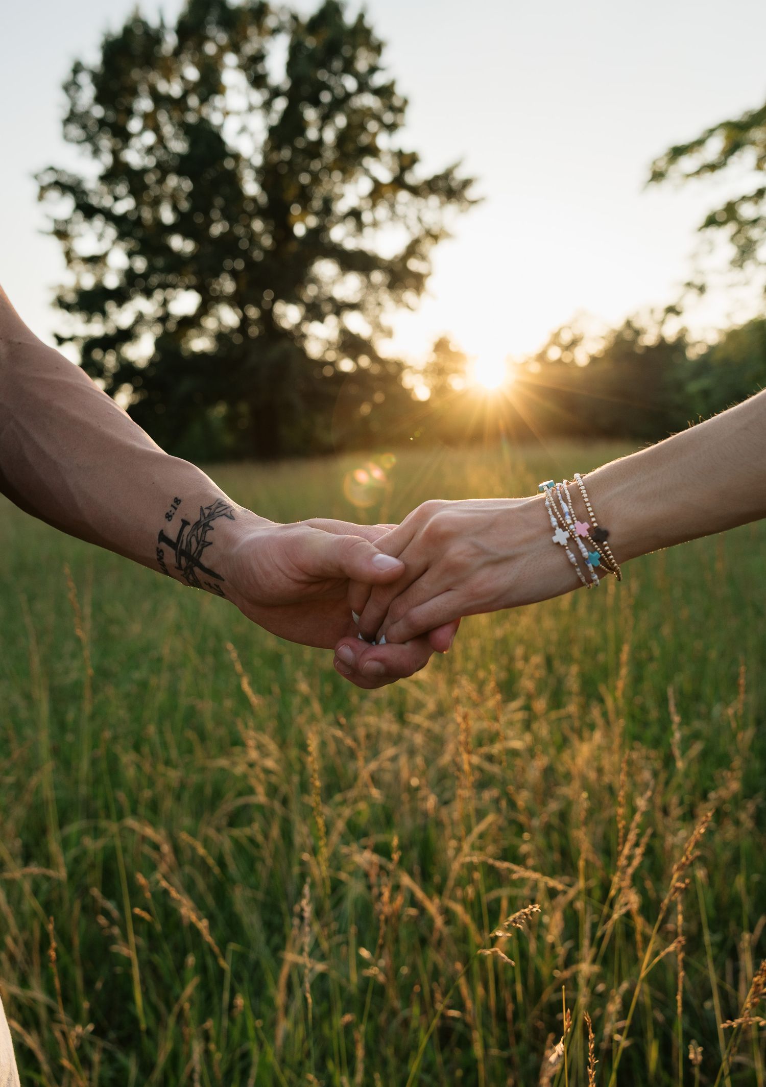 Couple holding hands in a sunlit field at sunset, one with a tattoo and the other wearing bracelets.
