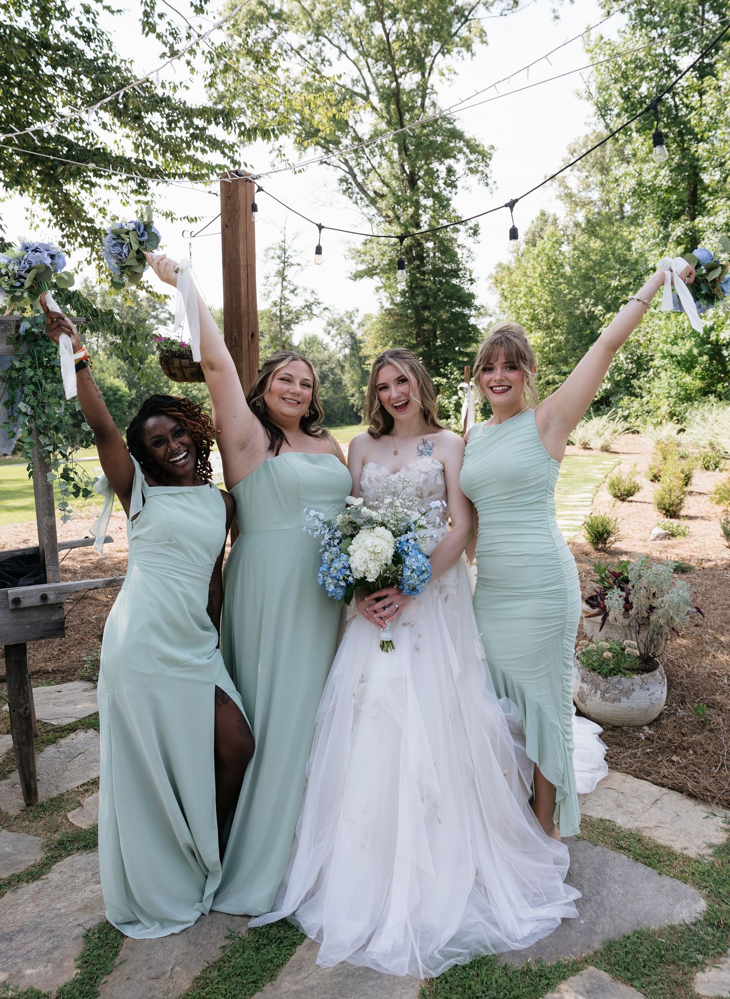Bride in white gown celebrating with three bridesmaids in sage green dresses at outdoor wedding.