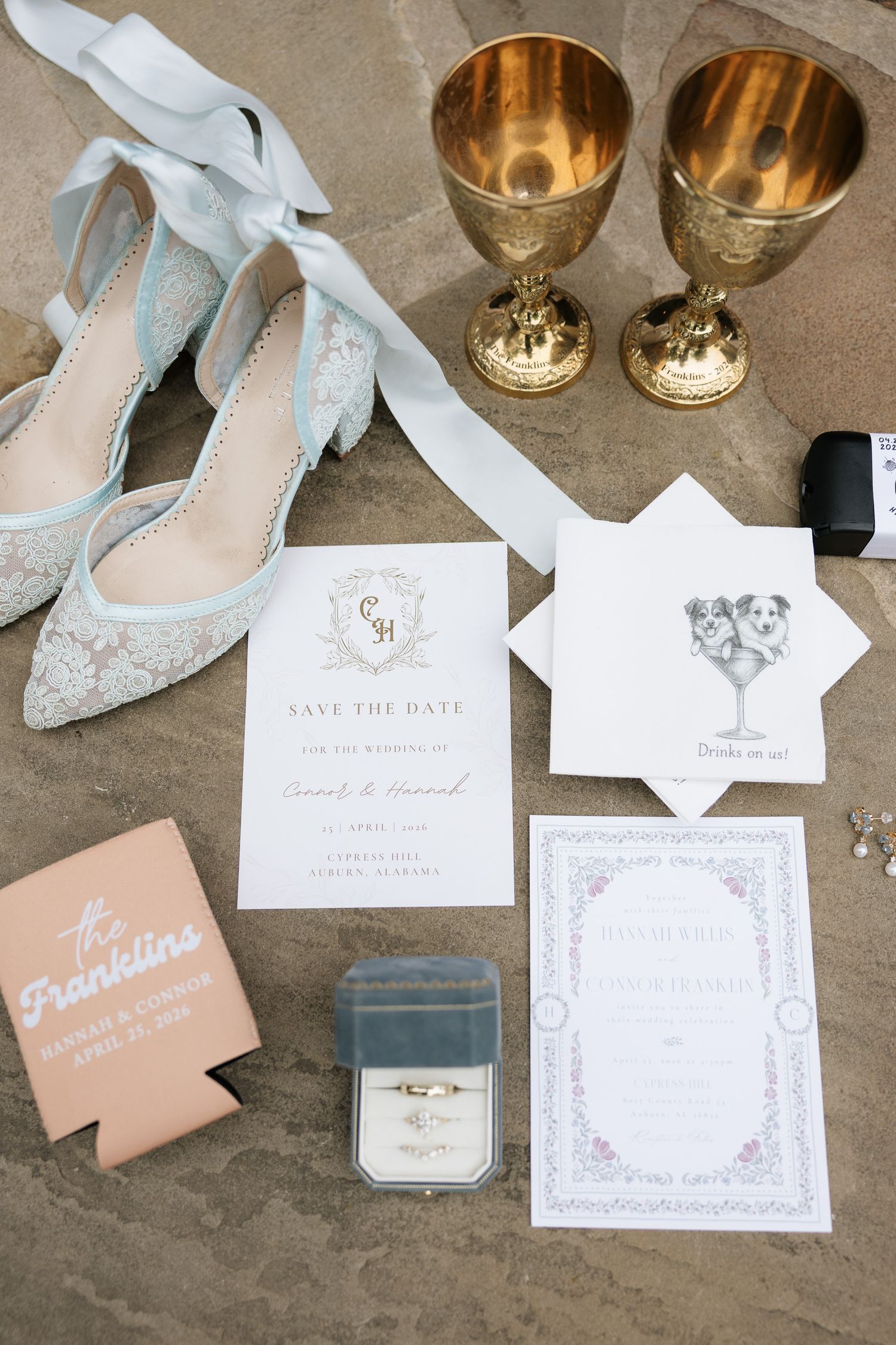 Elegant wedding flat lay with lace heels, gold goblets, save the date cards, ring box, and personalized koozie on stone surface.