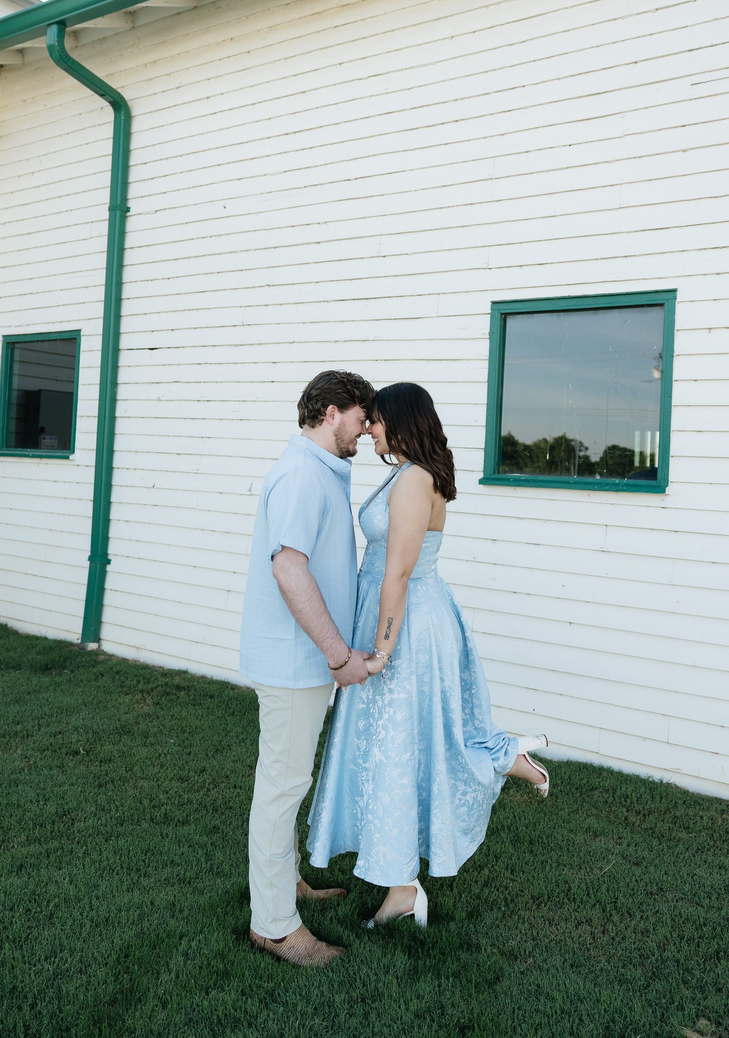 Couple in matching light blue outfits share a romantic moment outside a white building with green trim on a grassy lawn.