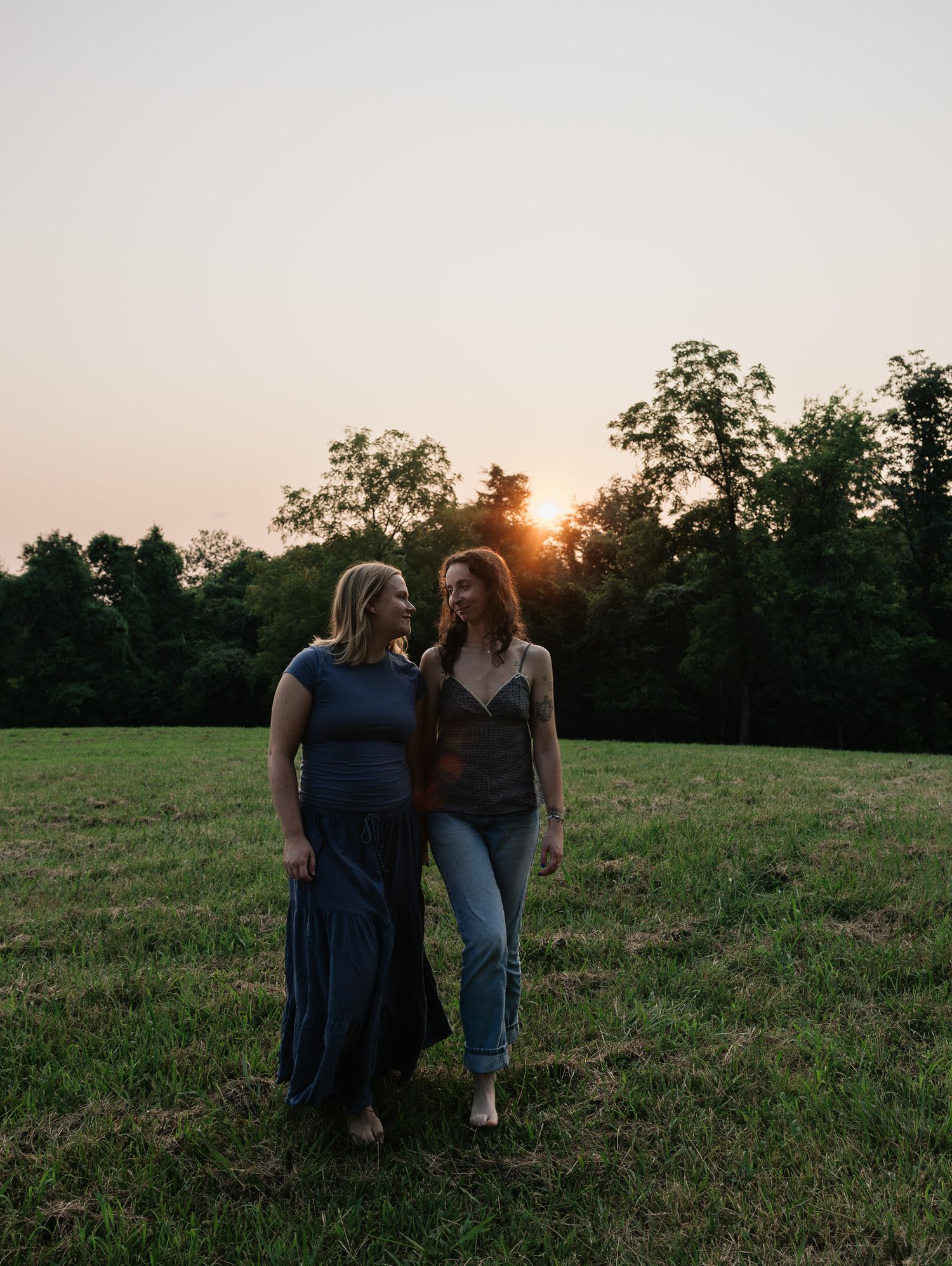 Two women walk together in a grassy field at golden hour sunset with trees in background.