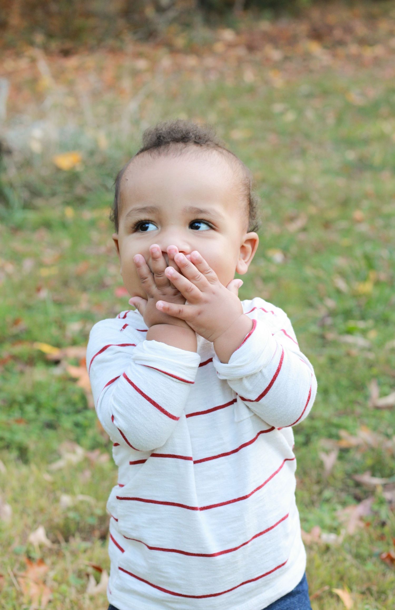 Baby in striped white and red shirt covering mouth with hands while looking up.