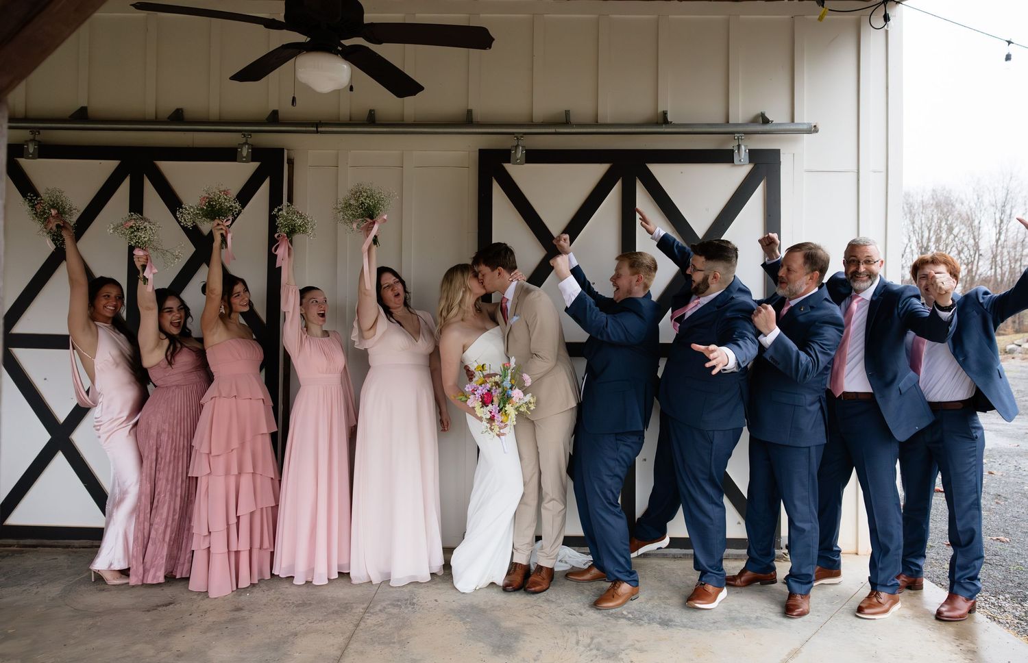 Wedding party celebrating outside barn doors, bridesmaids in pink gowns and groomsmen in navy suits raising hands joyfully.