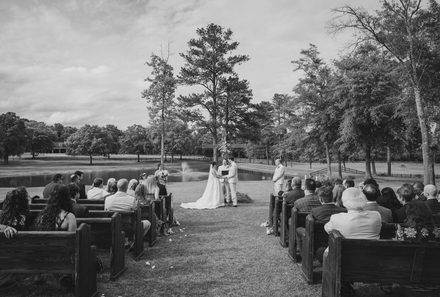 Black and white outdoor wedding ceremony with guests seated on wooden pews, couple at altar near a scenic pond and trees.
