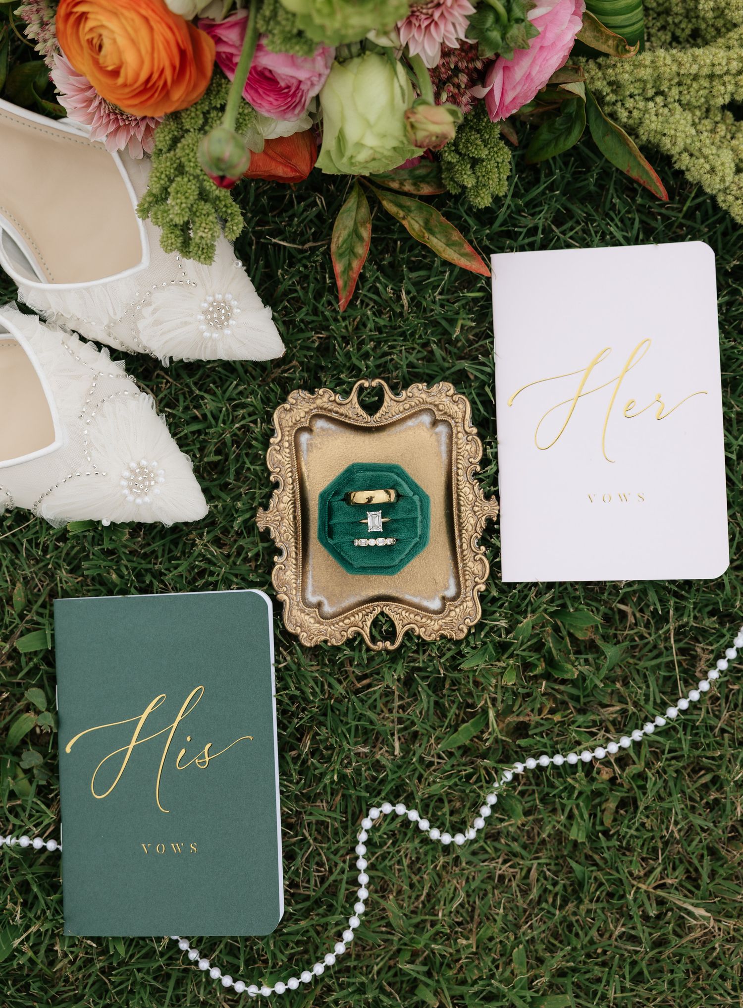 Wedding flat lay with His and Hers vow books, gold ring box, pearl necklace, bridal heels, and colorful floral bouquet on grass.
