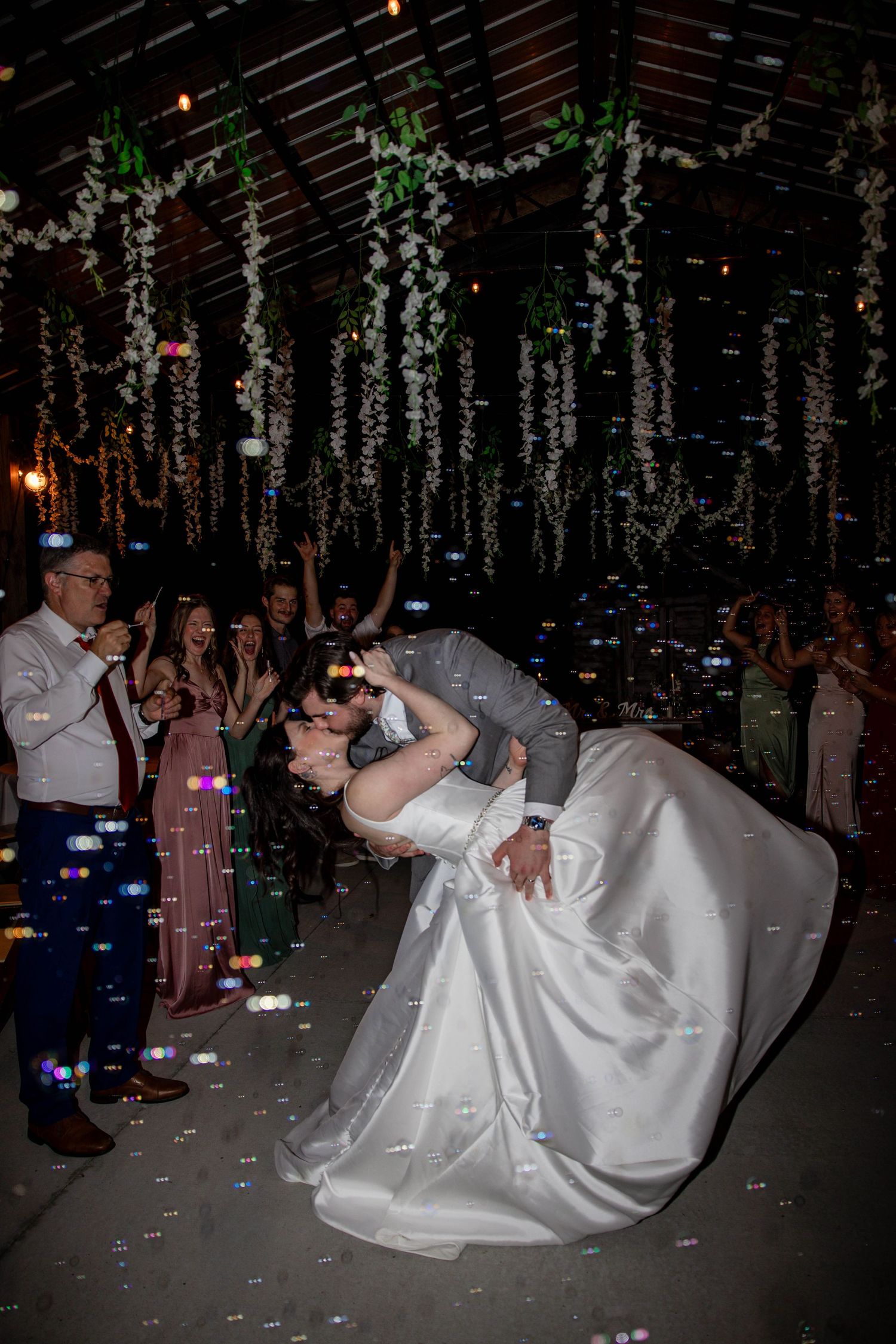Wedding reception features dramatic dip dance move under hanging floral decorations and string lights.