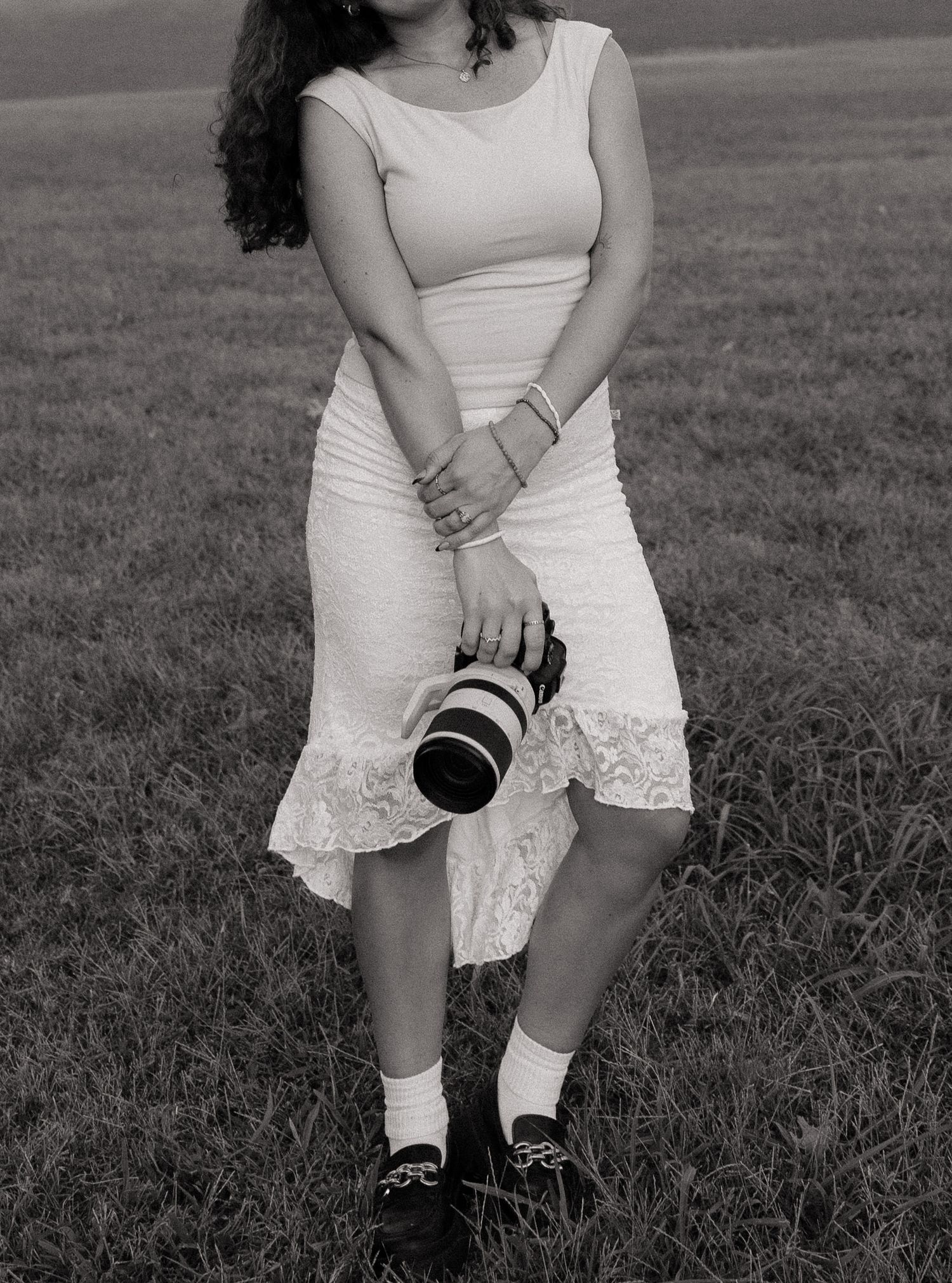 Black and white photo of a woman in a white lace dress holding a DSLR camera while standing in a grassy field.