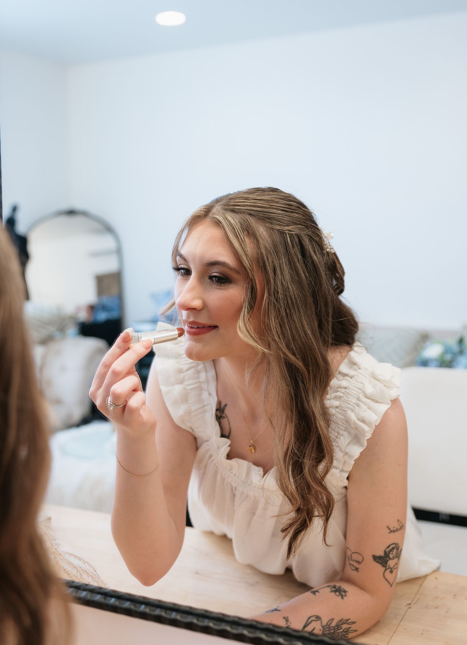 Bride applying lipstick while looking in a mirror, wearing a white ruffled dress with tattoos visible.