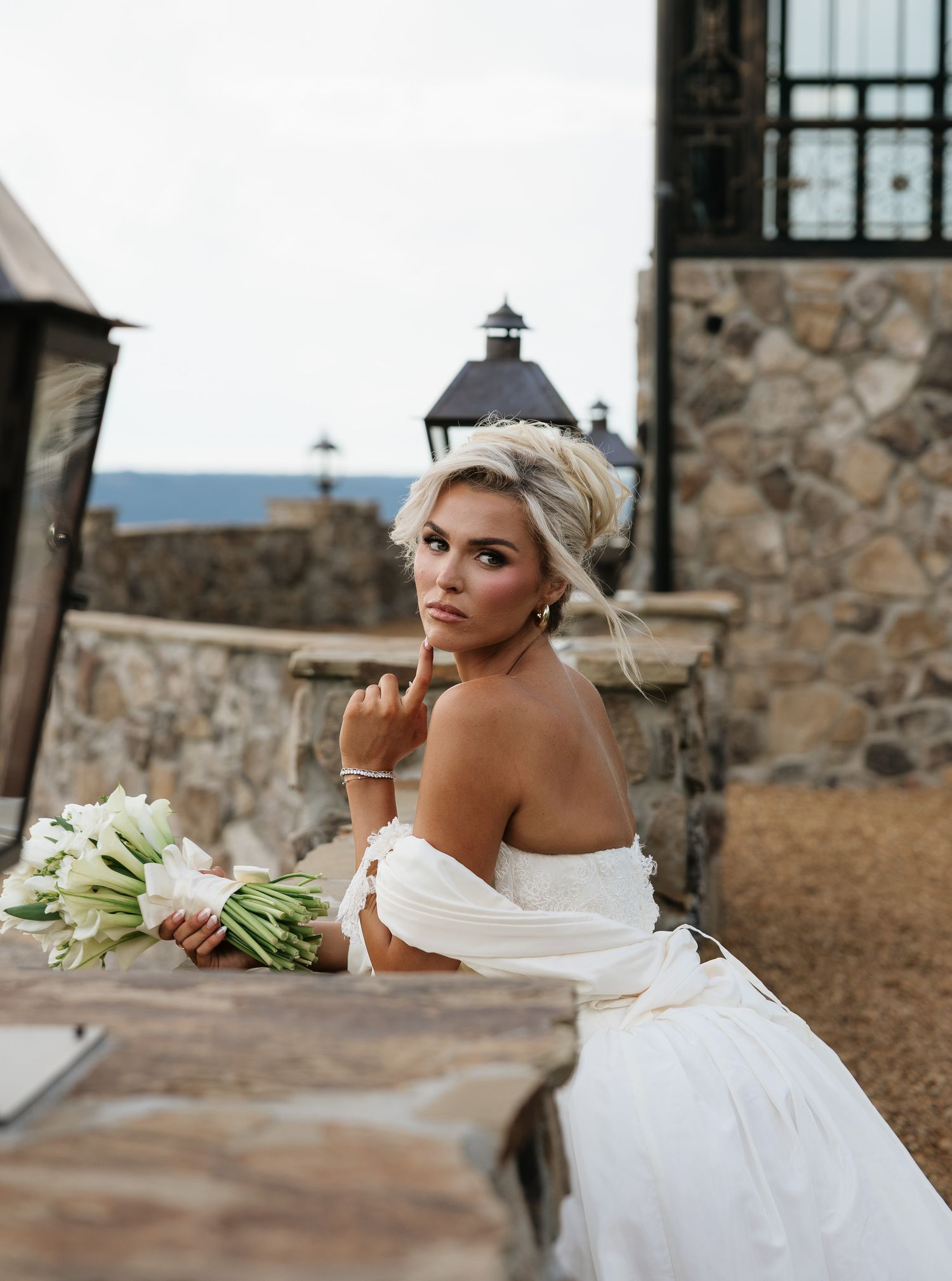 Elegant bride in off-shoulder white gown holding calla lily bouquet, seated at stone table at rustic outdoor venue.
