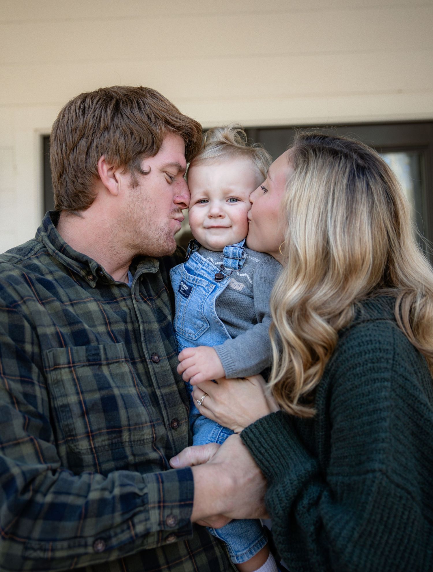 A couple kisses their smiling toddler on the cheeks in a heartwarming family portrait outdoors.