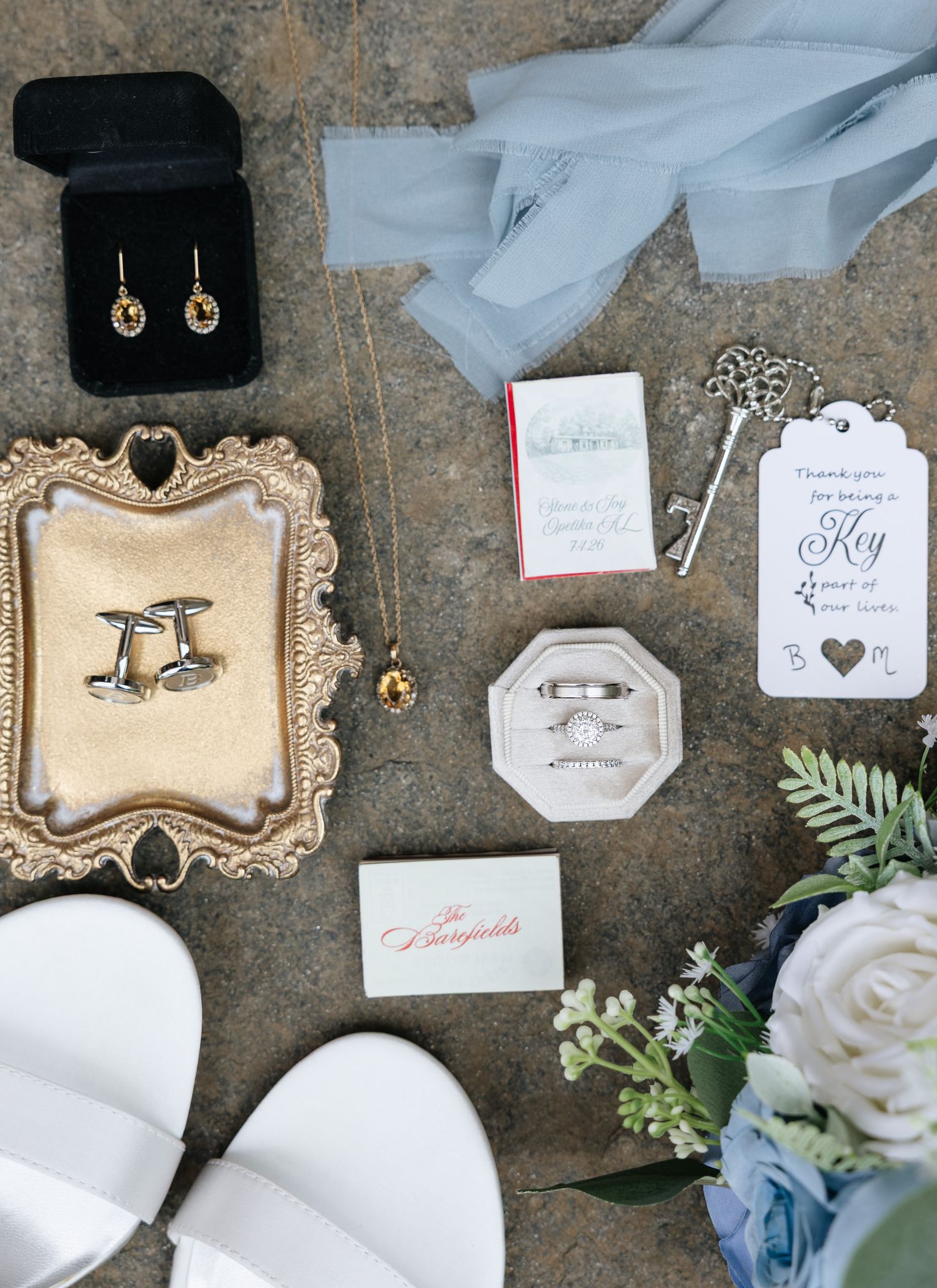Flat lay of wedding details including rings, cufflinks, earrings, bouquet, and accessories on stone surface.