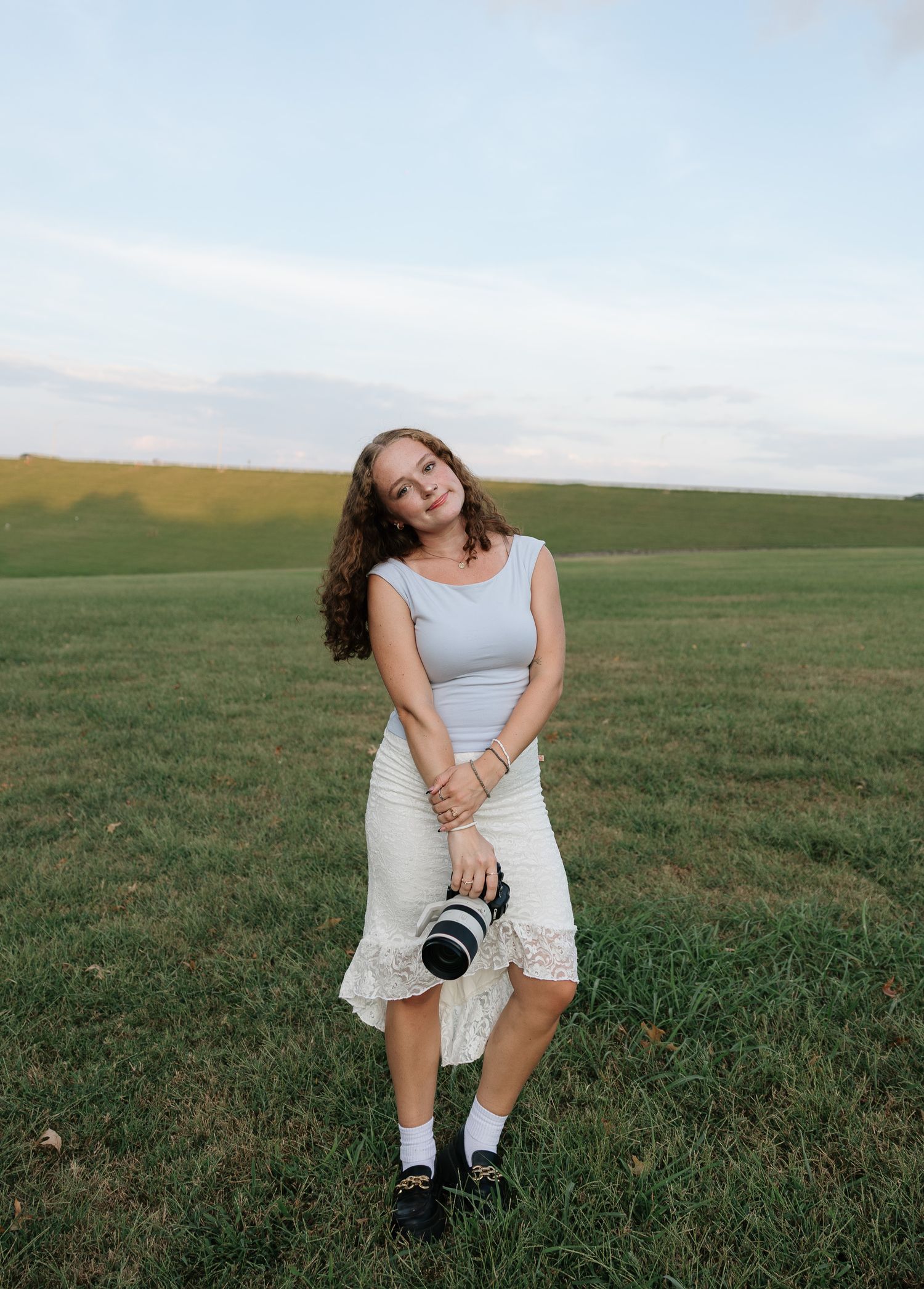 Young woman in white skirt and gray top holds camera lens while standing in green field under cloudy sky.
