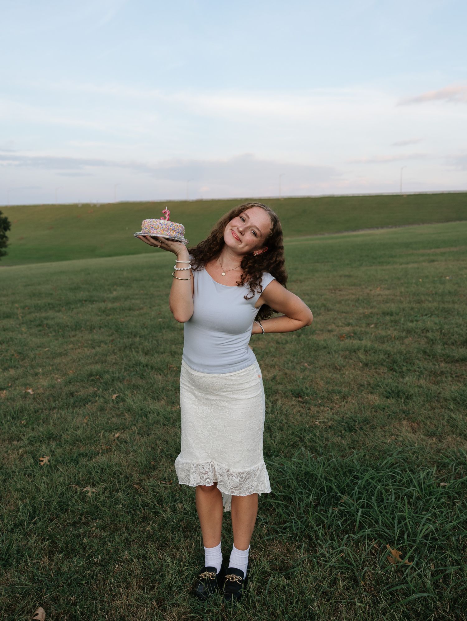 Young woman smiling outdoors holding a birthday cake in a green field, wearing a blue top and white lace skirt.