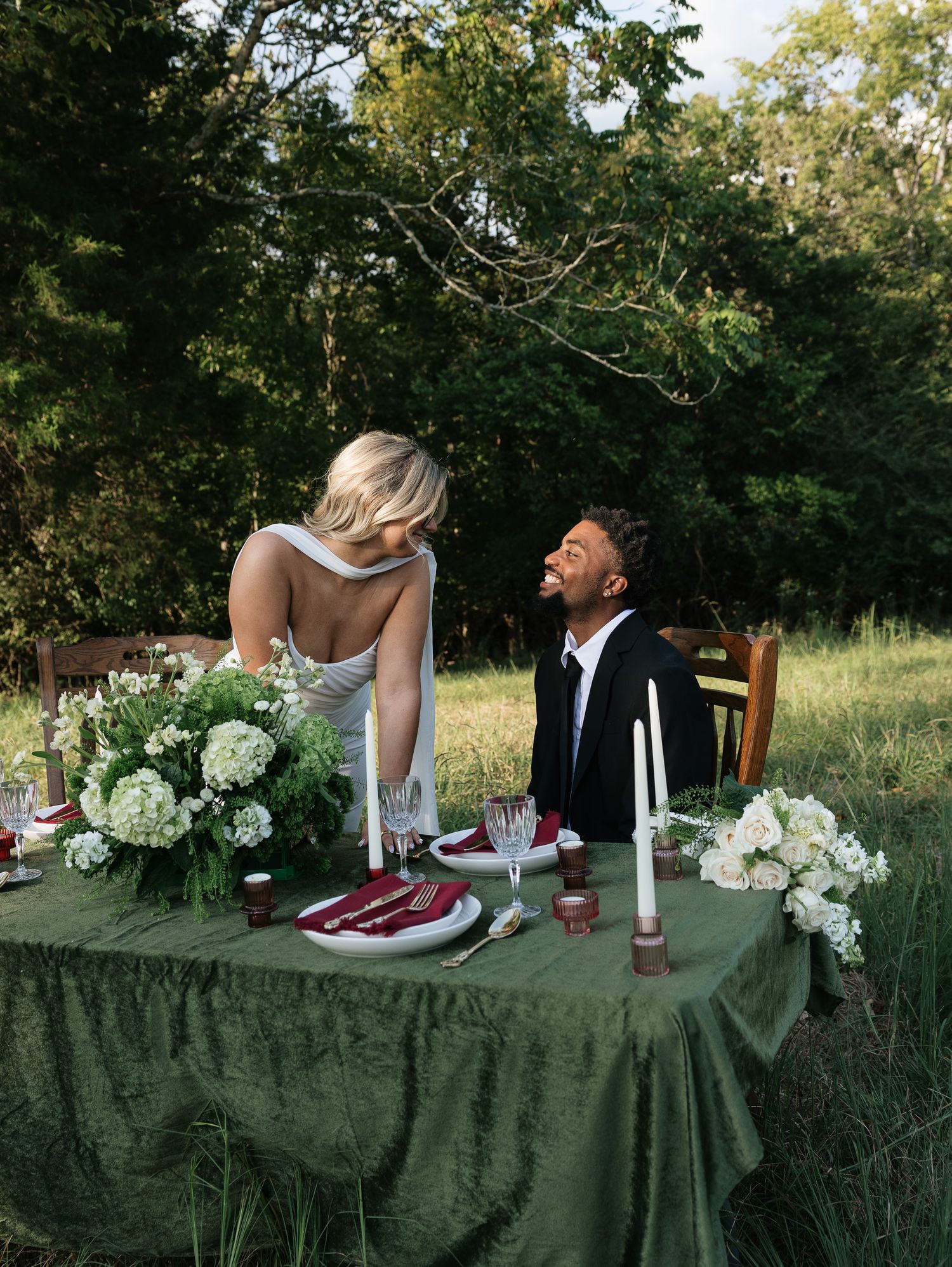 Couple shares a joyful moment at an elegant outdoor wedding table with green velvet cloth and white floral arrangements.
