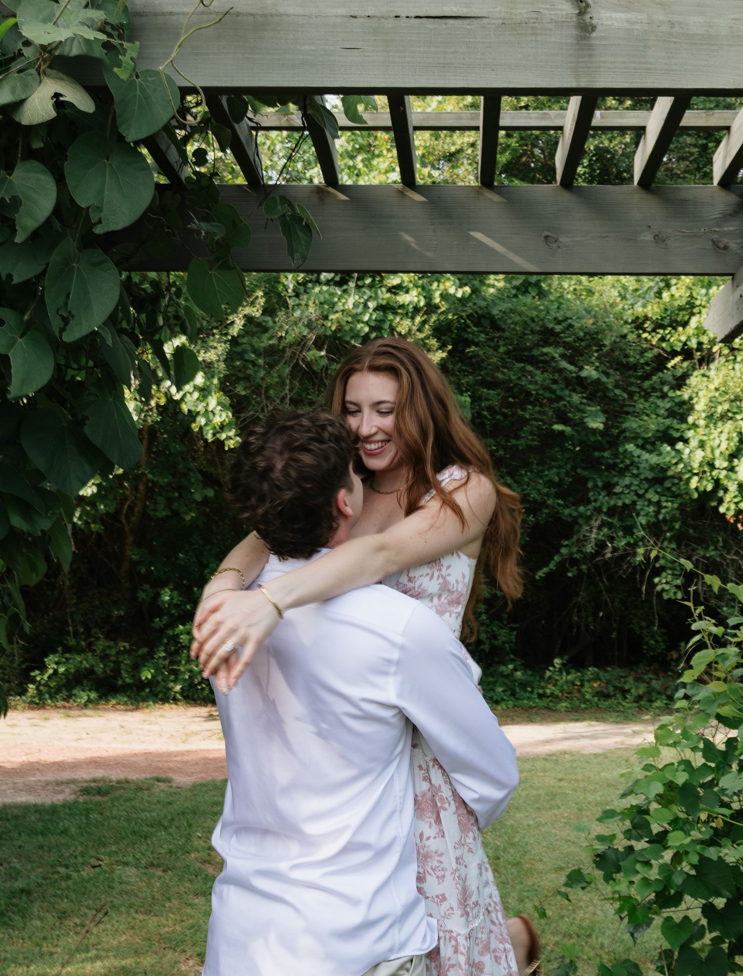 A joyful couple embraces and dances under a wooden pergola surrounded by lush green garden foliage.
