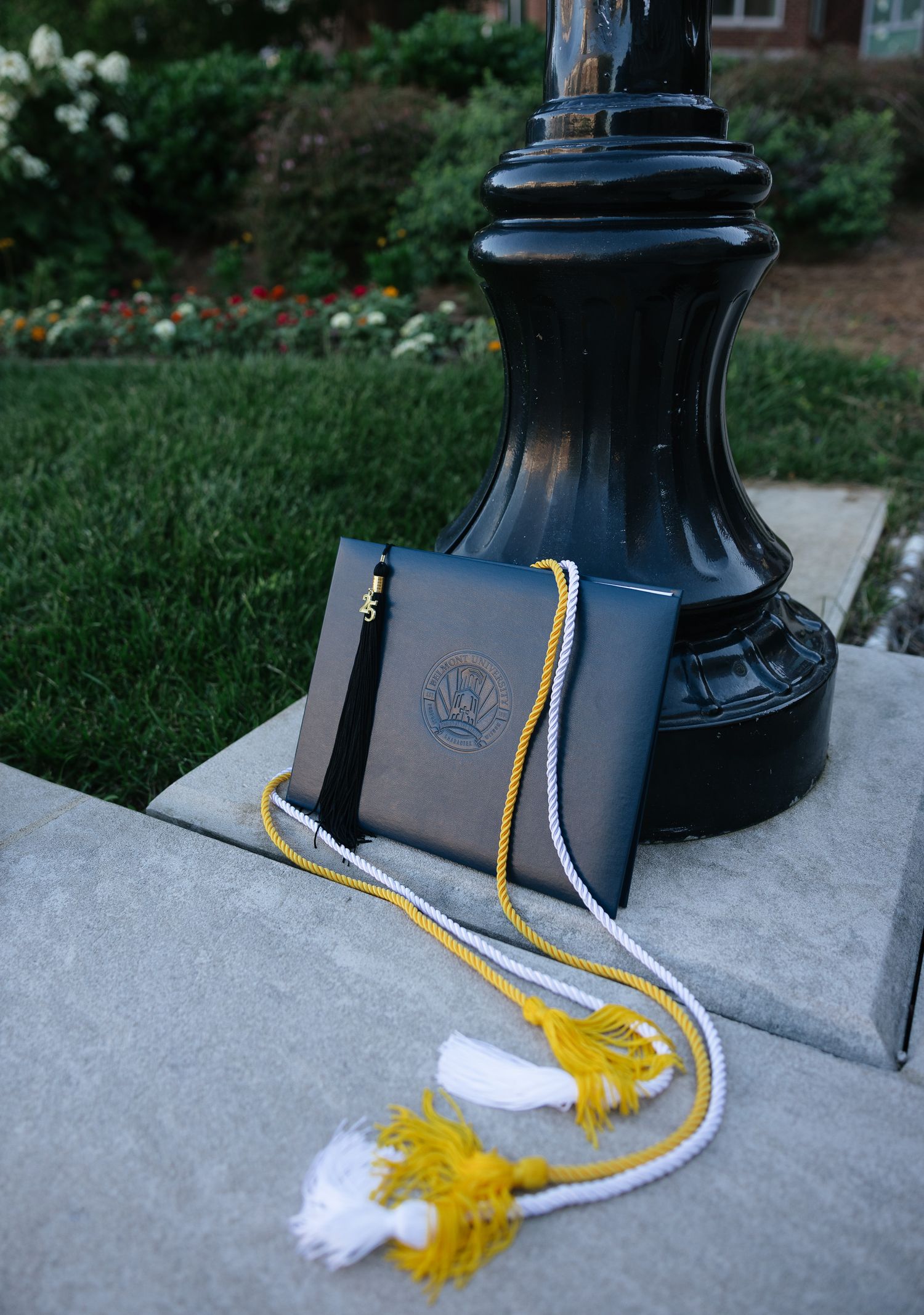 Graduation cap and honor cords with gold and white tassels displayed on stone pedestal outdoors.